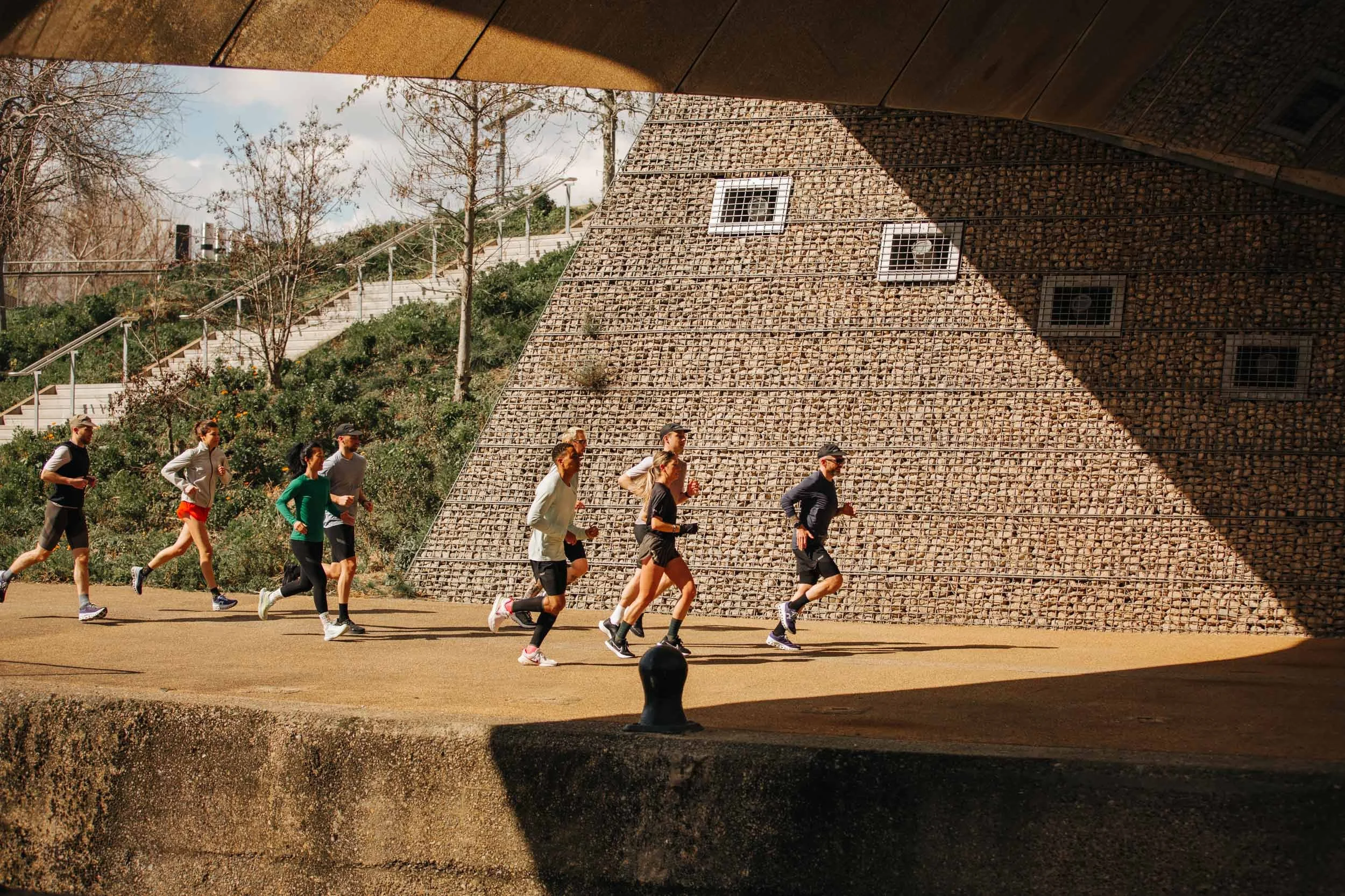 A group of people jogging outdoors on a path, with a sloped rock wall and trees in the background, seen from under a bridge or overhang.