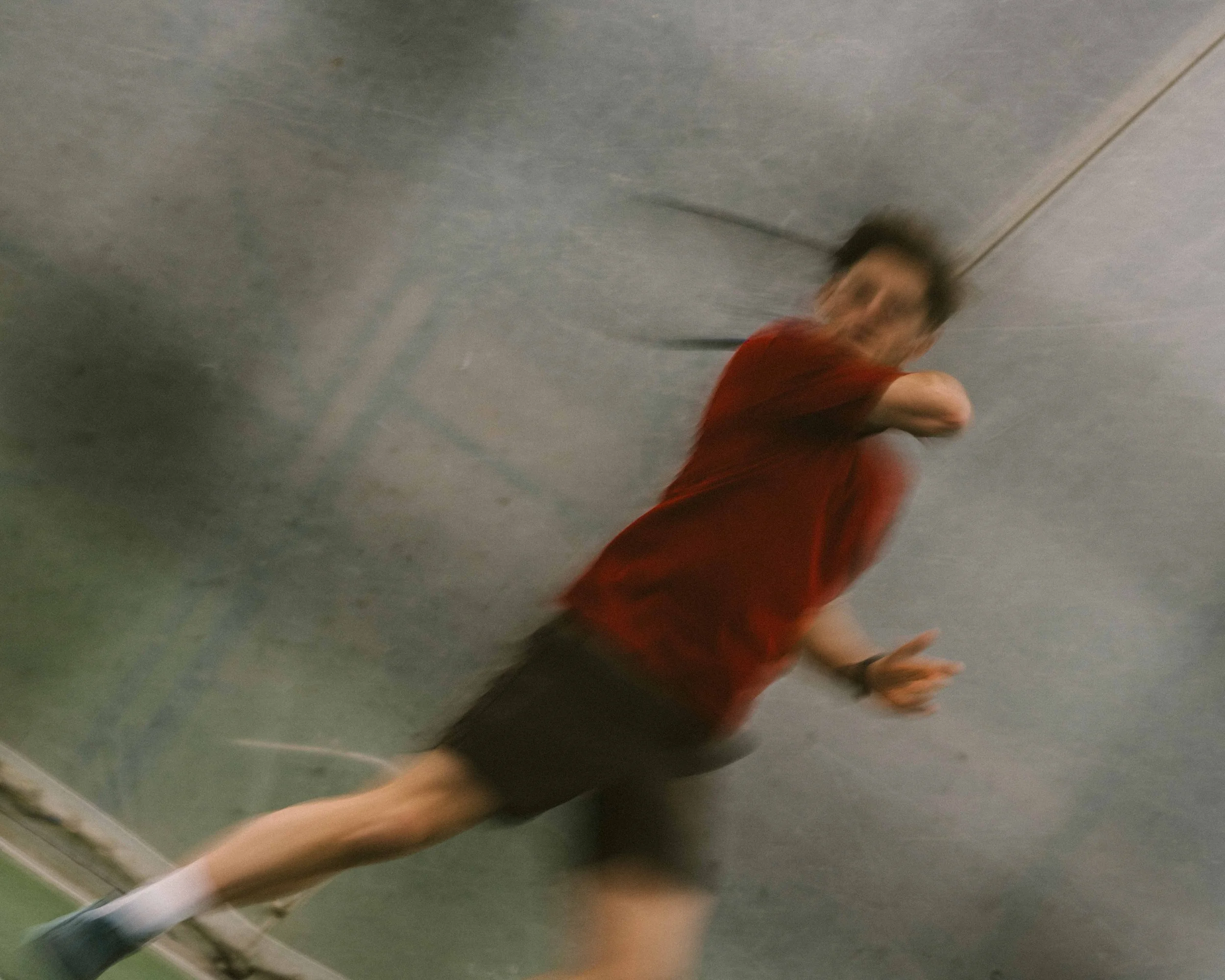 Motion-blurred photo of a person in a red shirt and black shorts falling or slipping on a gray floor, with an unclear facial expression.