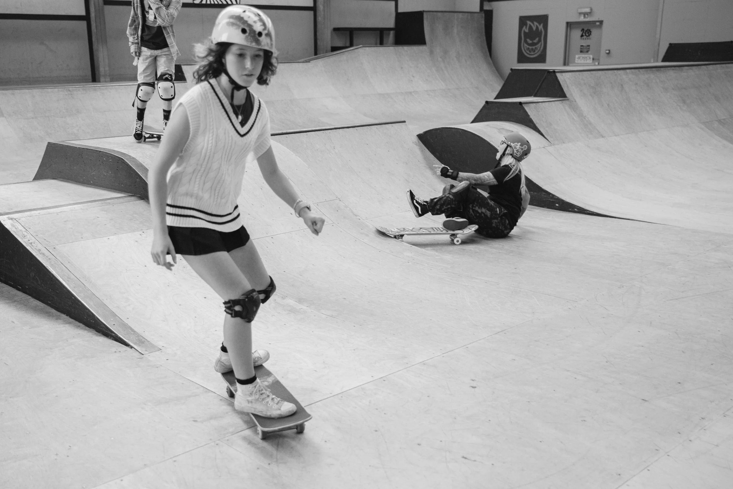 A girl riding a skateboard at an indoor skatepark, wearing a helmet, shorts, a sleeveless sweater, and knee pads, with another girl in the background sitting on the floor with a skateboard, also wearing a helmet.