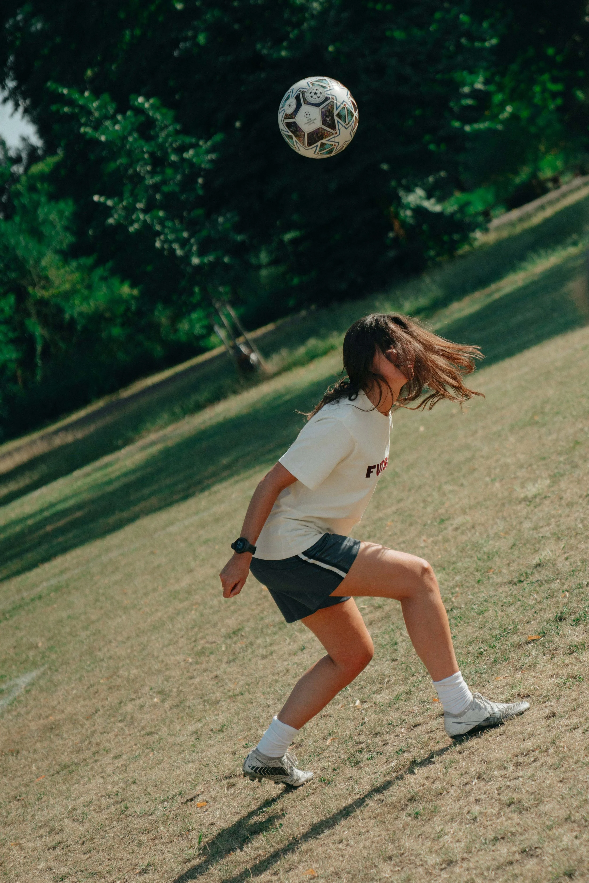 A girl playing soccer outdoors, kicking a patterned soccer ball in a grassy park with trees in the background.