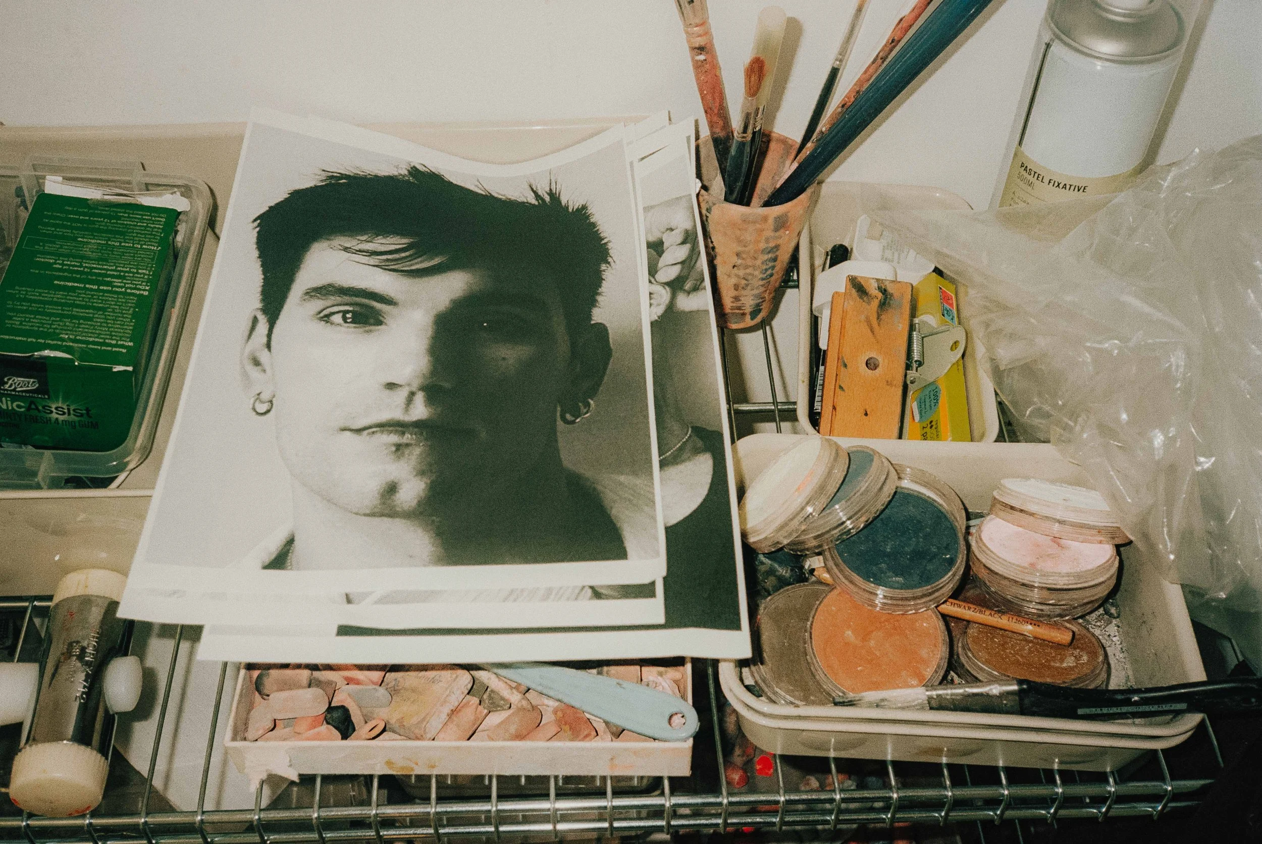 A cluttered desk with black and white photographs of a young man, brushes, jars of makeup, and various art supplies.