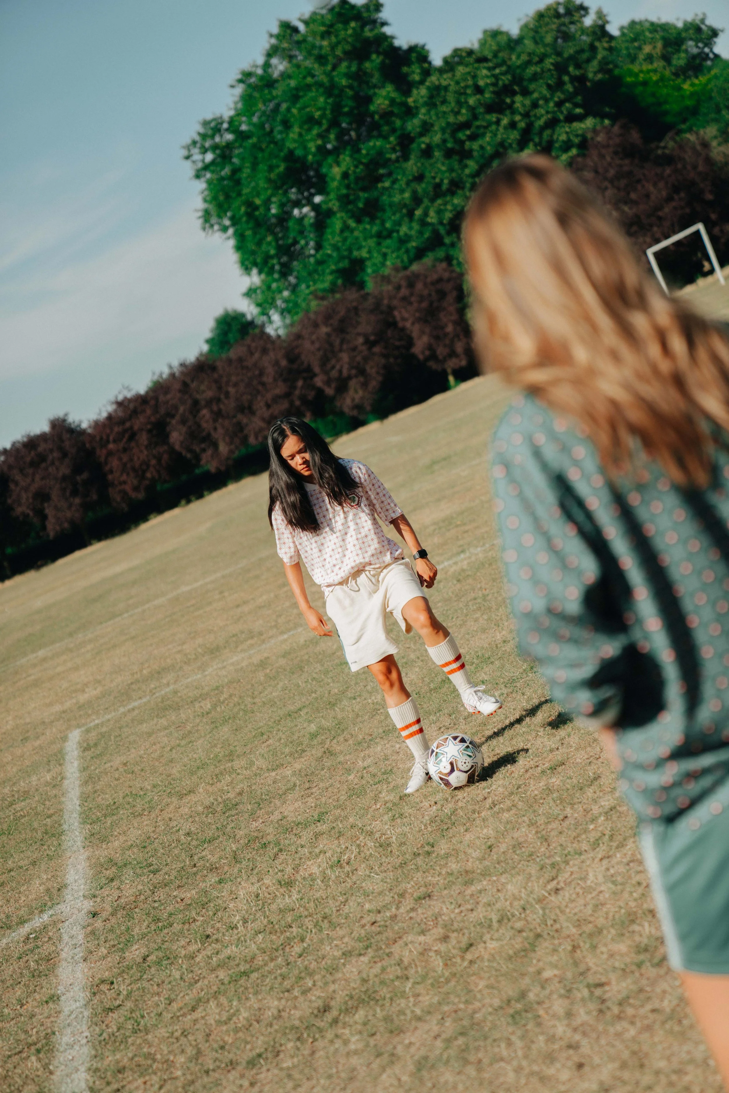 Two women playing soccer on a grassy field, with a woman in a patterned jersey about to kick the ball and another woman in the foreground, out of focus, with trees in the background.