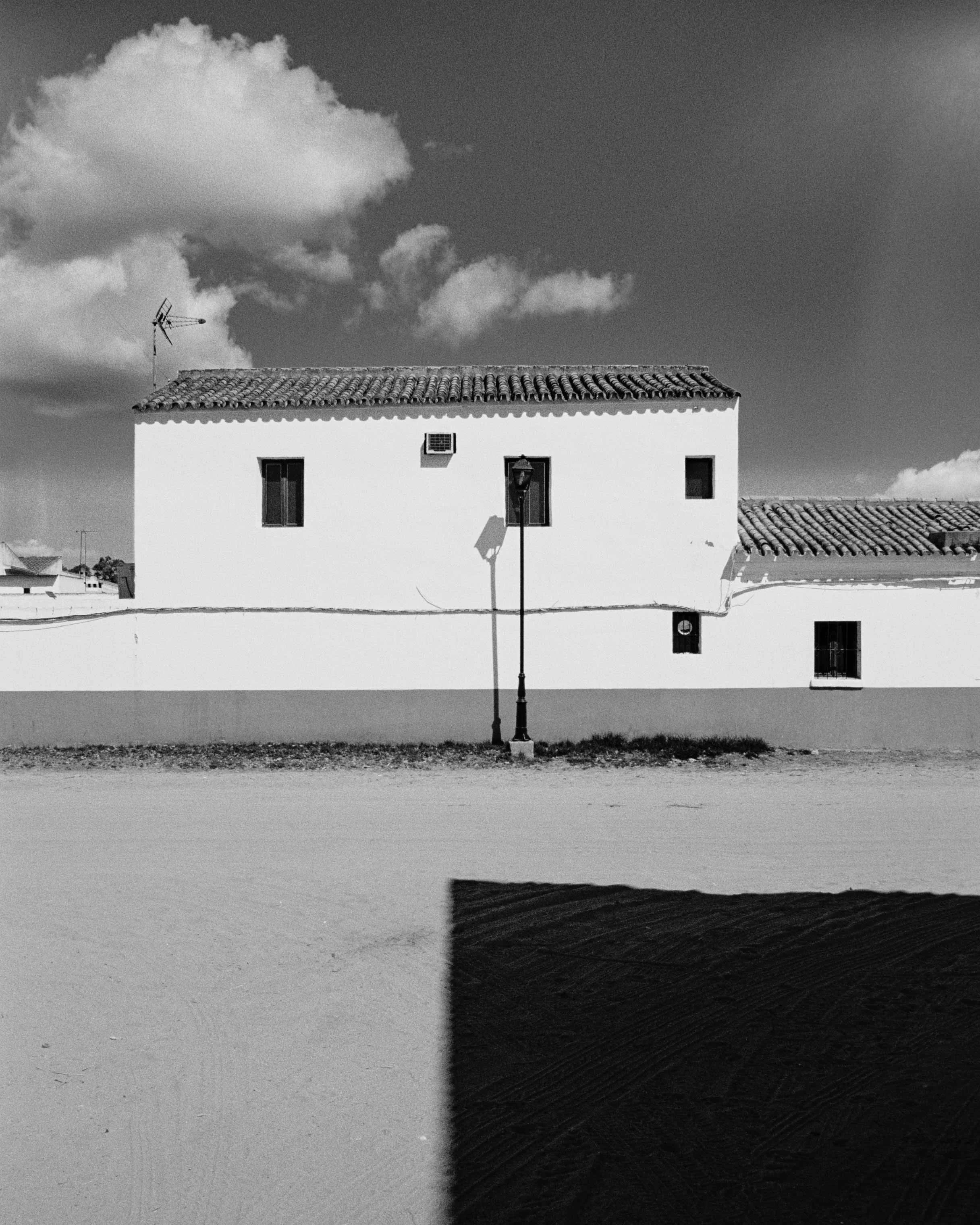 Black and white photo of a white building with a tiled roof, three windows, and a street lamp in the foreground, under a partly cloudy sky.