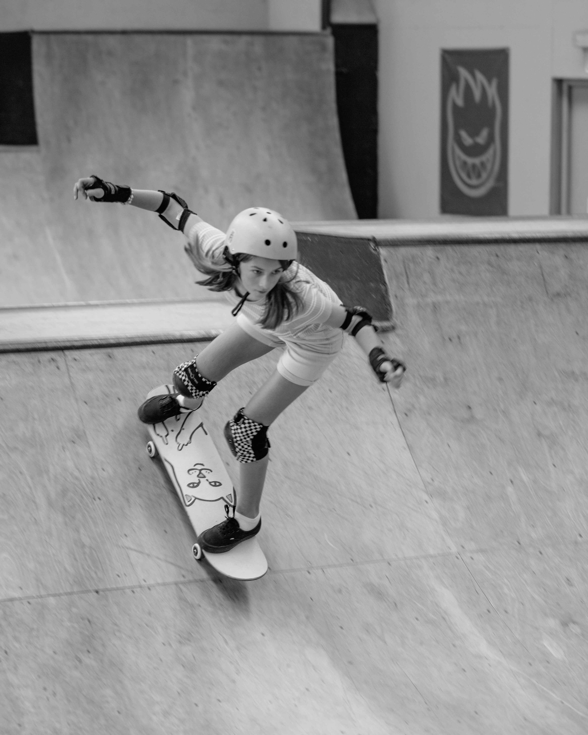 A young girl skateboarding at an indoor skatepark, wearing a helmet, protective pads, and checkered knee pads.