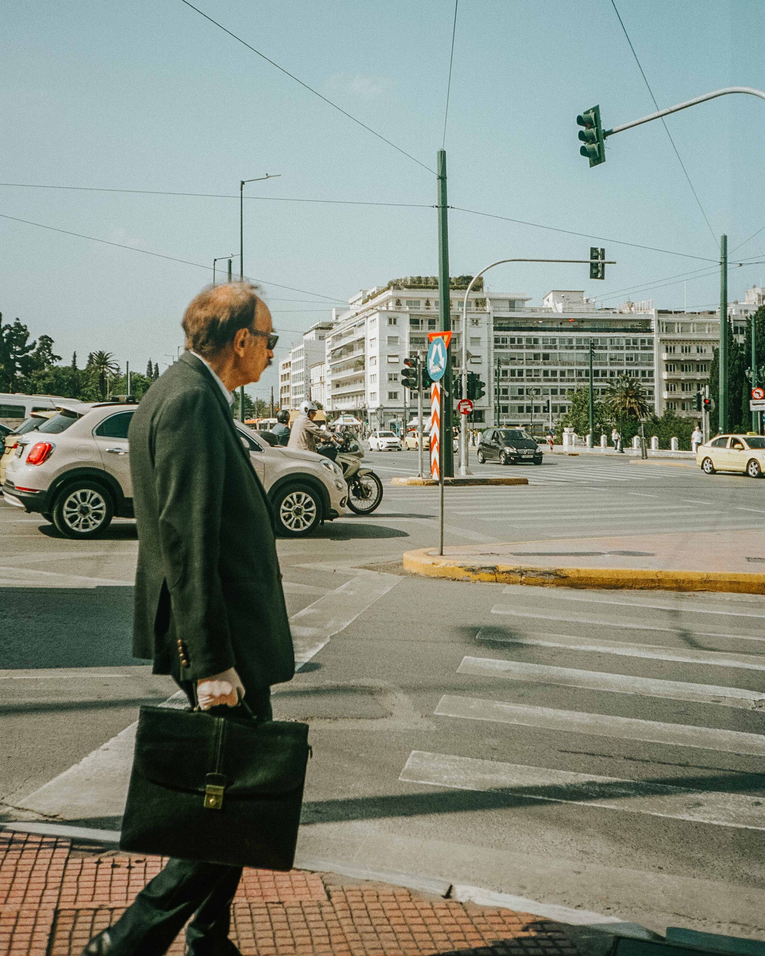 A man wearing a suit and sunglasses walking across a city street with a briefcase in hand, cars and motorcycles waiting at the traffic light, and buildings in the background under a clear sky.