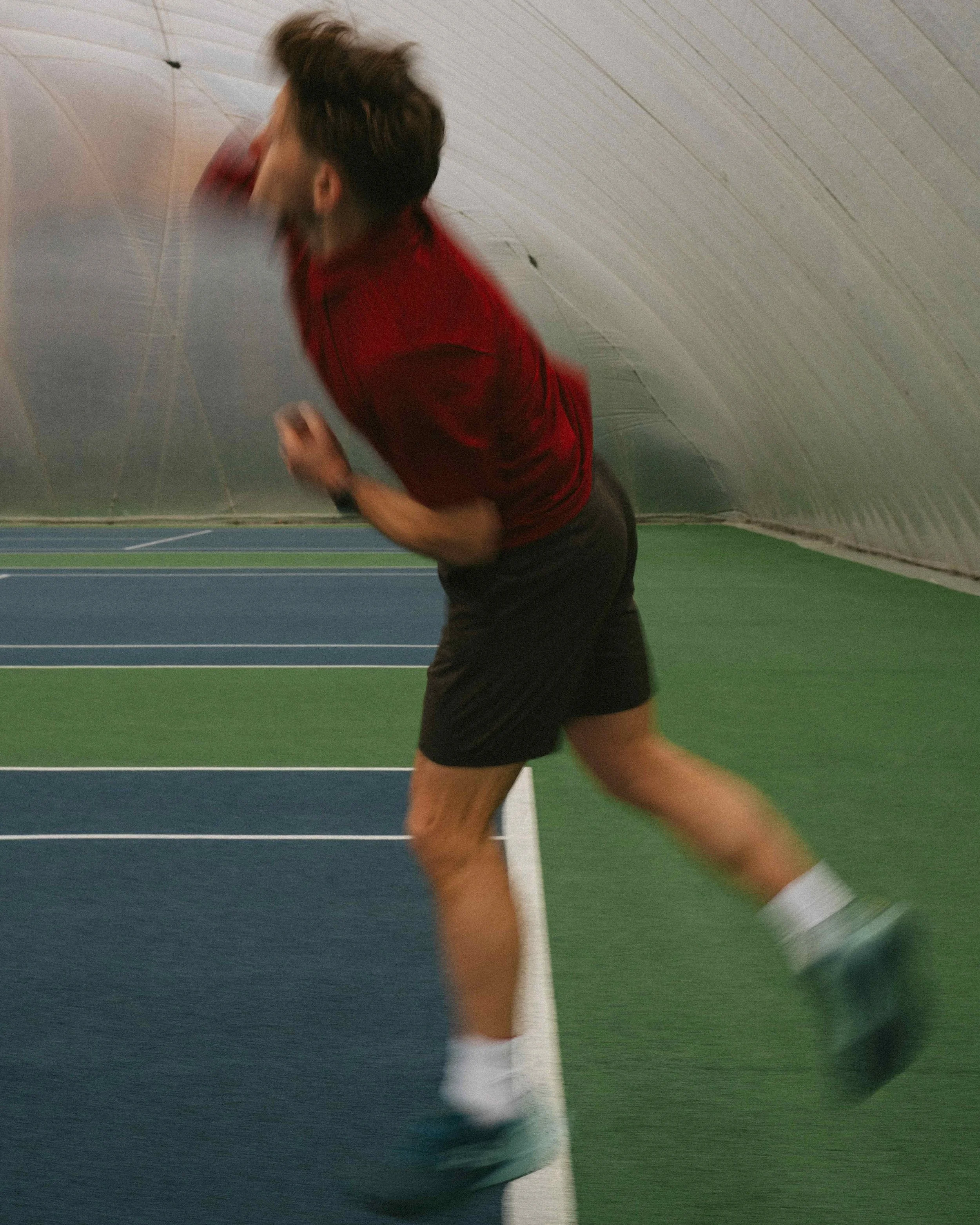 A man running indoors on a track inside a tunnel, wearing a red shirt, black shorts, and running shoes.