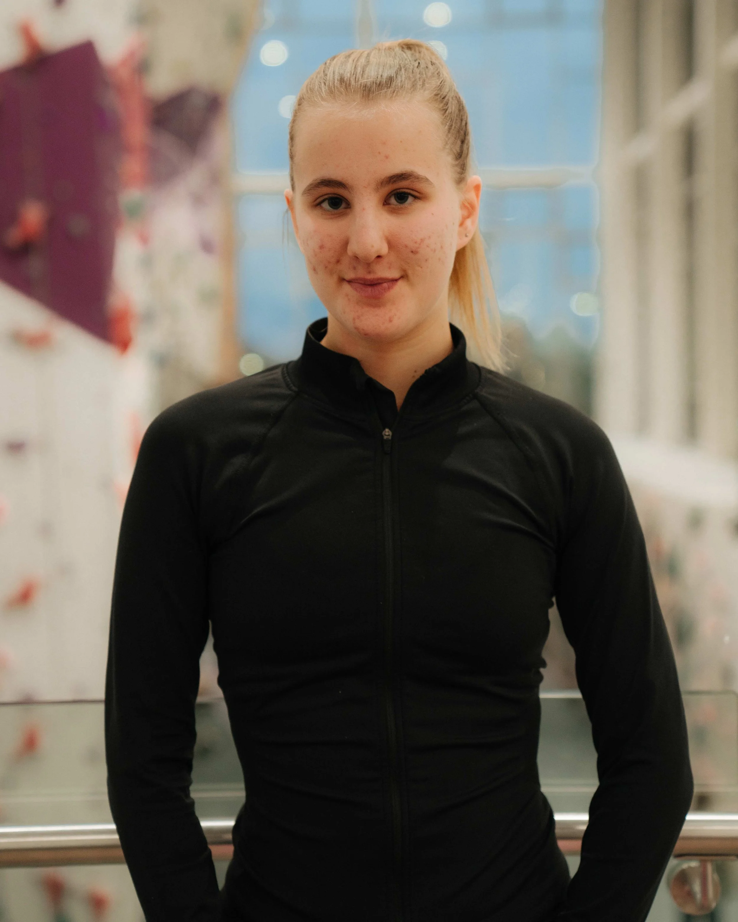 Young woman with blonde hair tied back, wearing a black athletic zip-up jacket, standing inside a building with a blurry background of colorful decorations and windows.