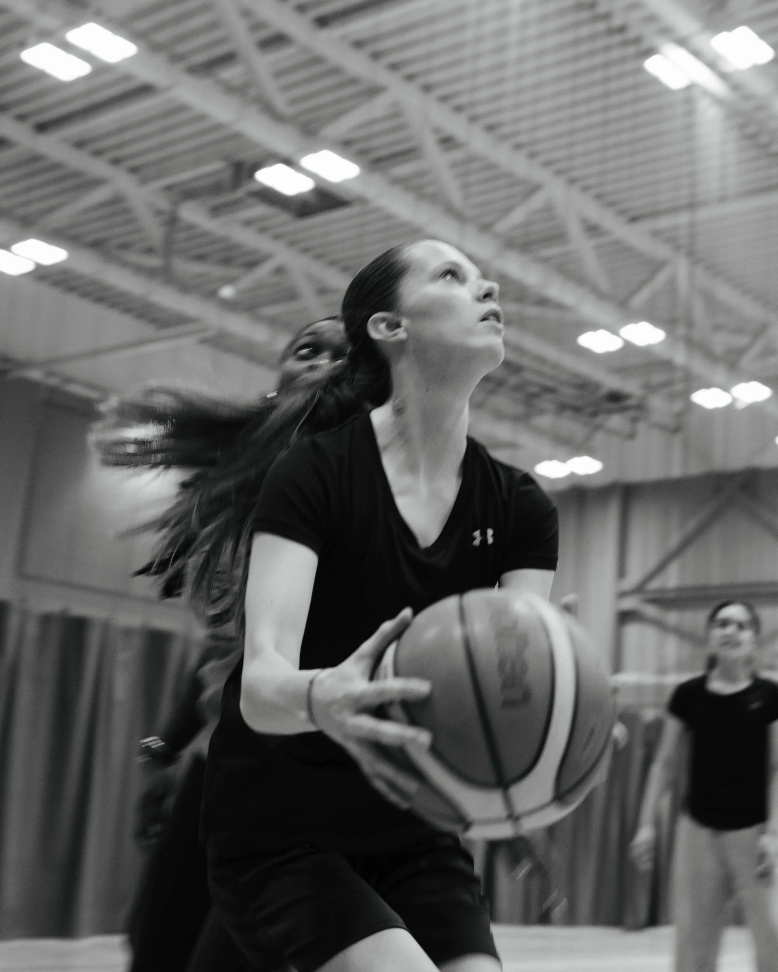 A black and white photo of women playing basketball indoors. One woman is holding a basketball, preparing to shoot, with her hair flowing behind her. There are other women in the background, and the gymnasium ceiling with lights is visible above.
