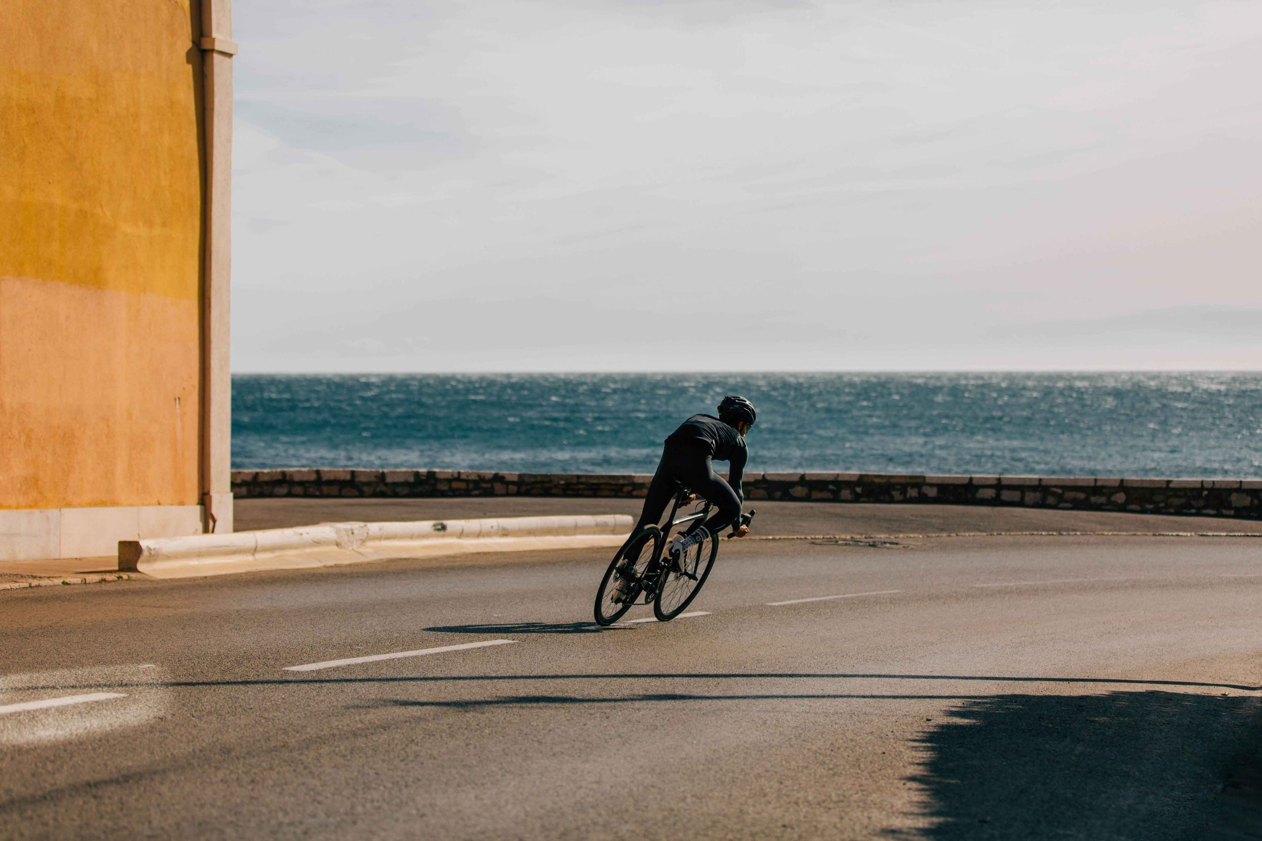 A person in black cycling gear riding a bicycle along a seaside road with the ocean in the background.