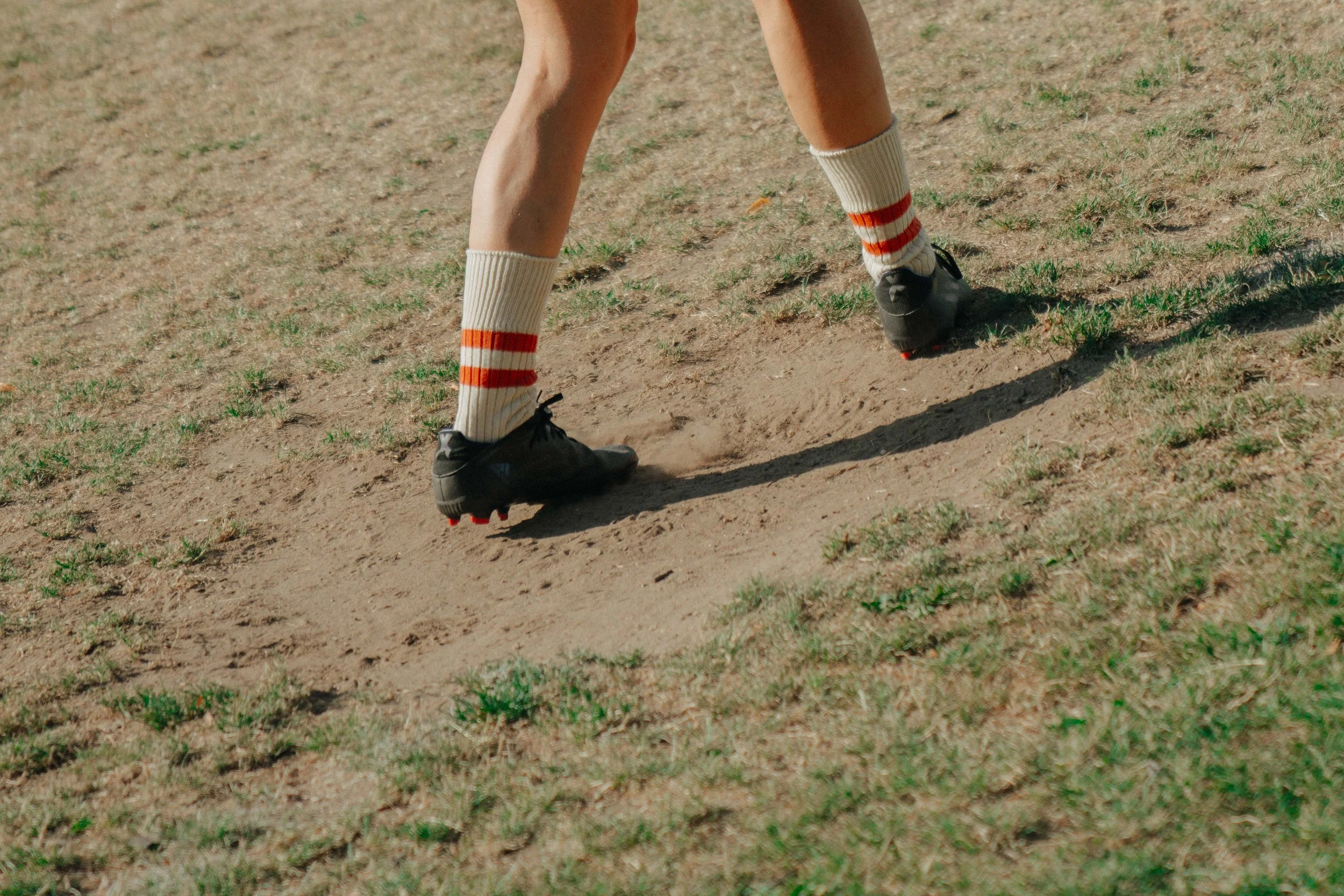 Close-up of a person's legs wearing striped knee-high socks and soccer cleats, playing soccer on a dirt field.