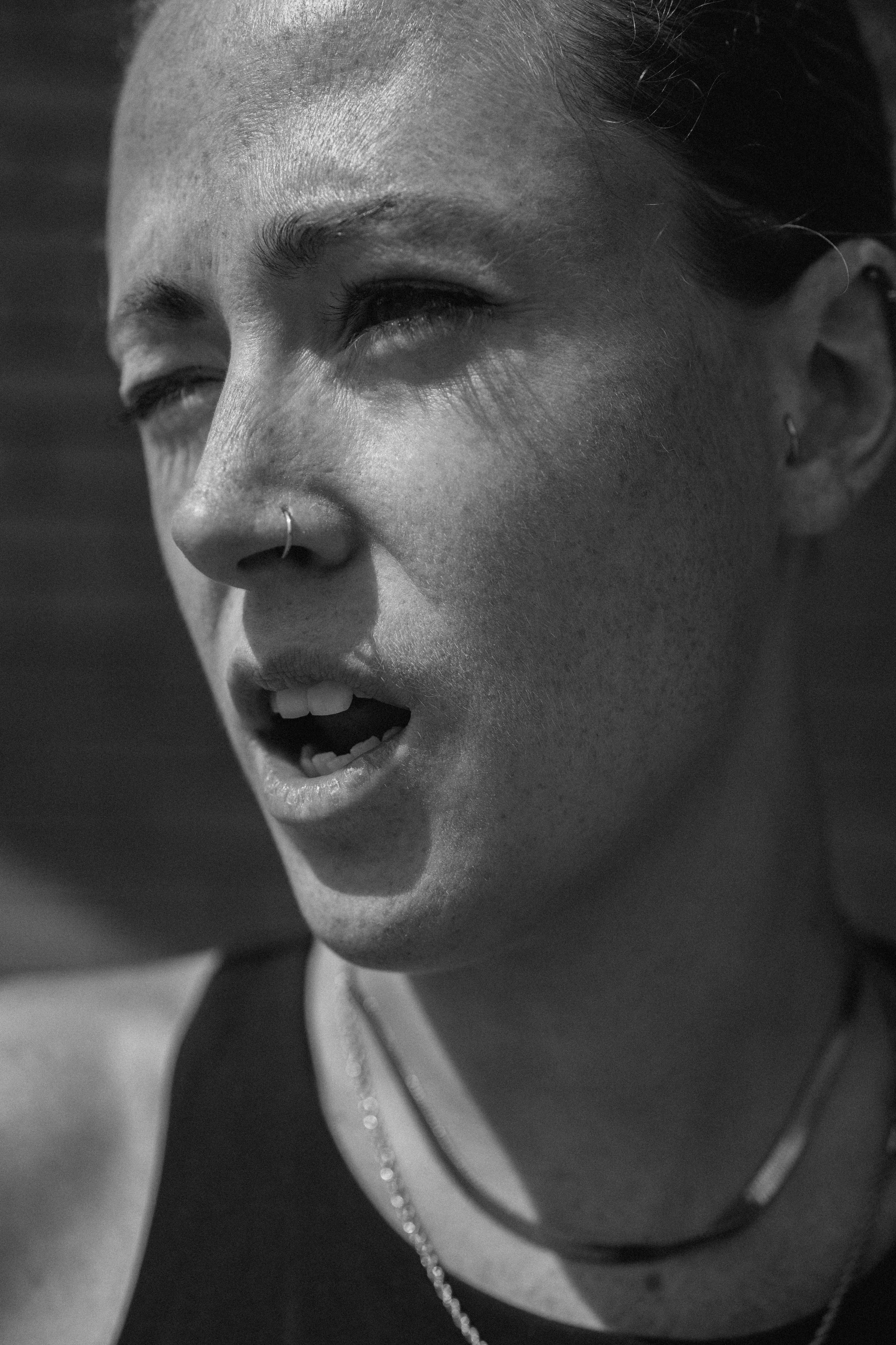 Close-up of a woman's face in black and white, with visible freckles, nose piercing, wearing necklaces, and her hair pulled back.