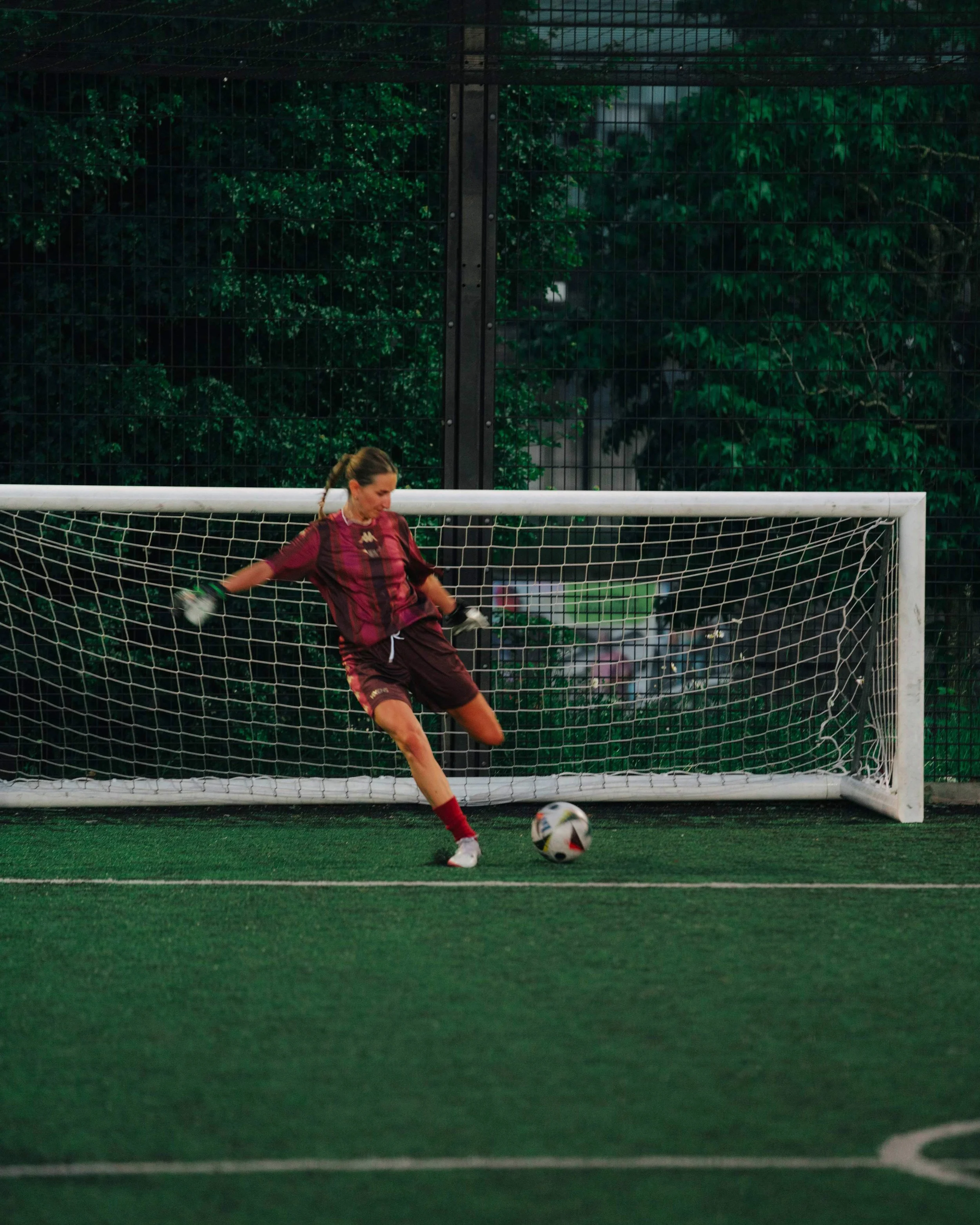 Female soccer goalie in maroon uniform kicking a soccer ball on a green field with a soccer net and green trees in the background.