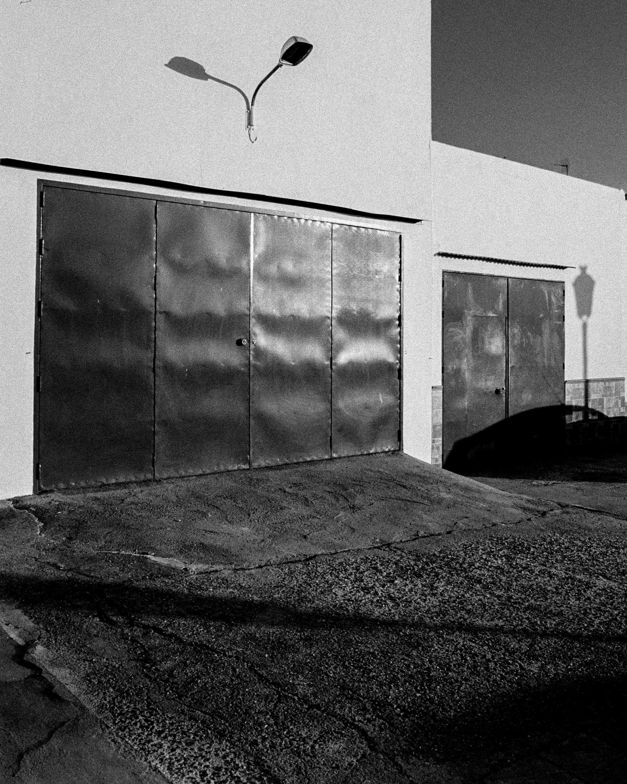Black and white photo of an industrial garage with metal doors, a street lamp, and a streetlight shadow on the building wall during nighttime.