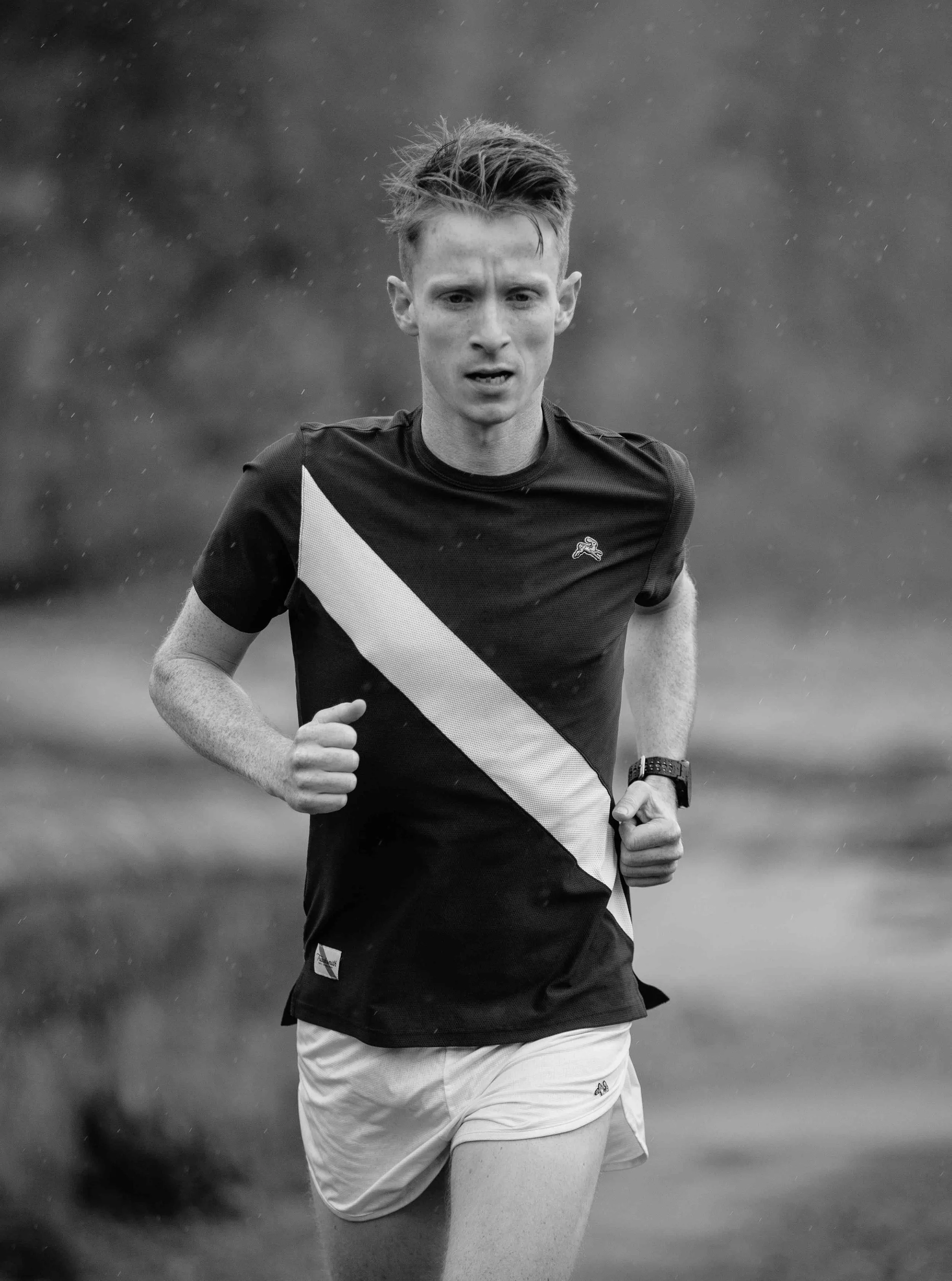 Black and white photo of a young man running outdoors, wearing a short-sleeved athletic shirt, shorts, and a watch, focused on his run.