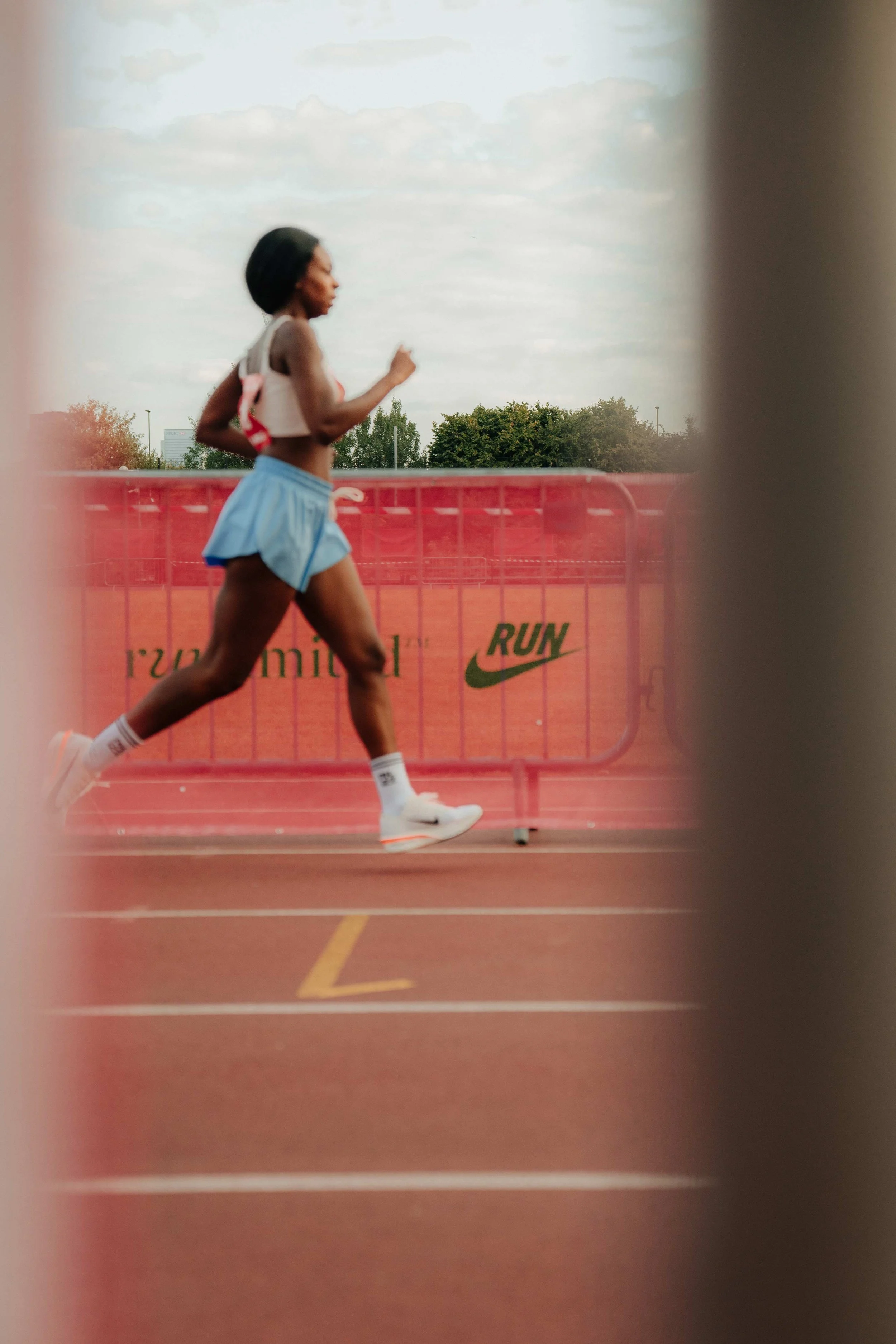 A female athlete running on a track seen through gaps in red barriers with Nike logos.