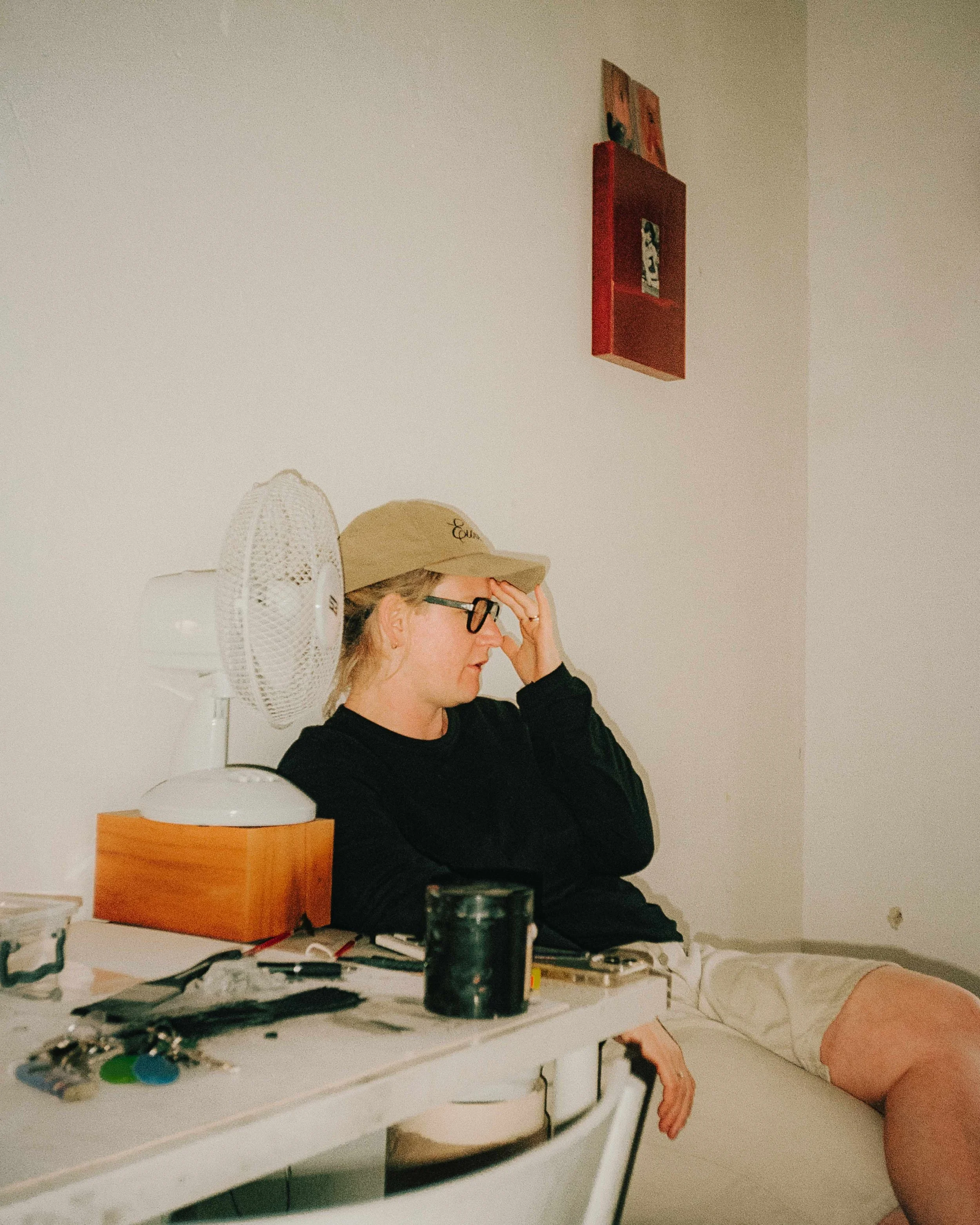 A woman wearing glasses and a beige cap sitting at a cluttered white table, resting her head on her hand with a distressed expression. Items on the table include a fan, keys, keys, and some electronics. There is a white wall and a red shelf hanging on it in the background.