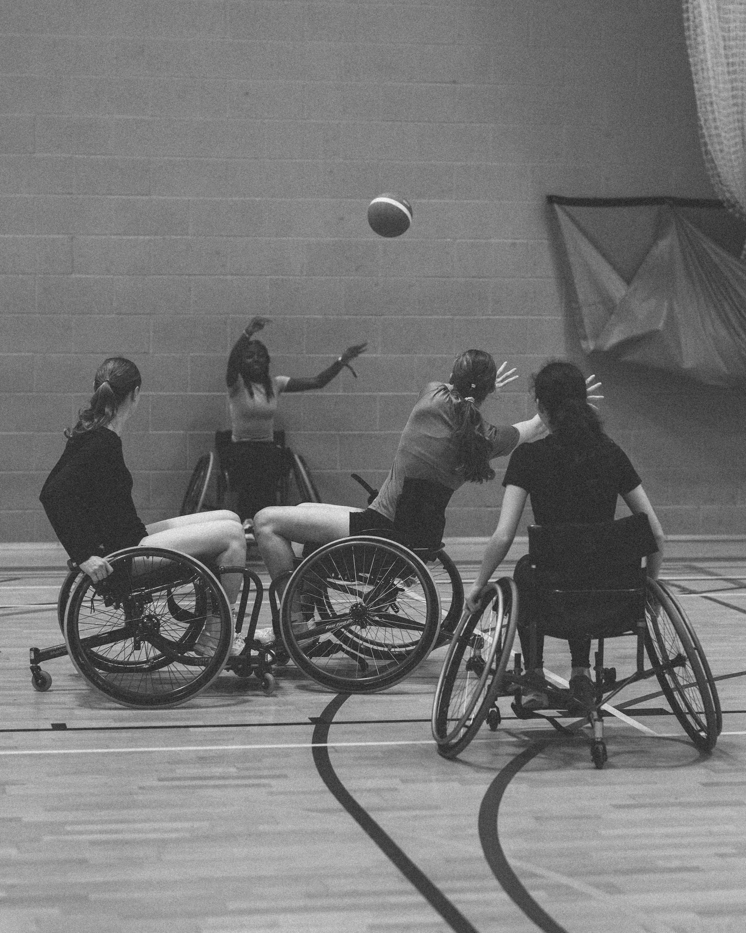 Four women in wheelchairs playing basketball indoors with a ball in the air and a volleyball net in the background.