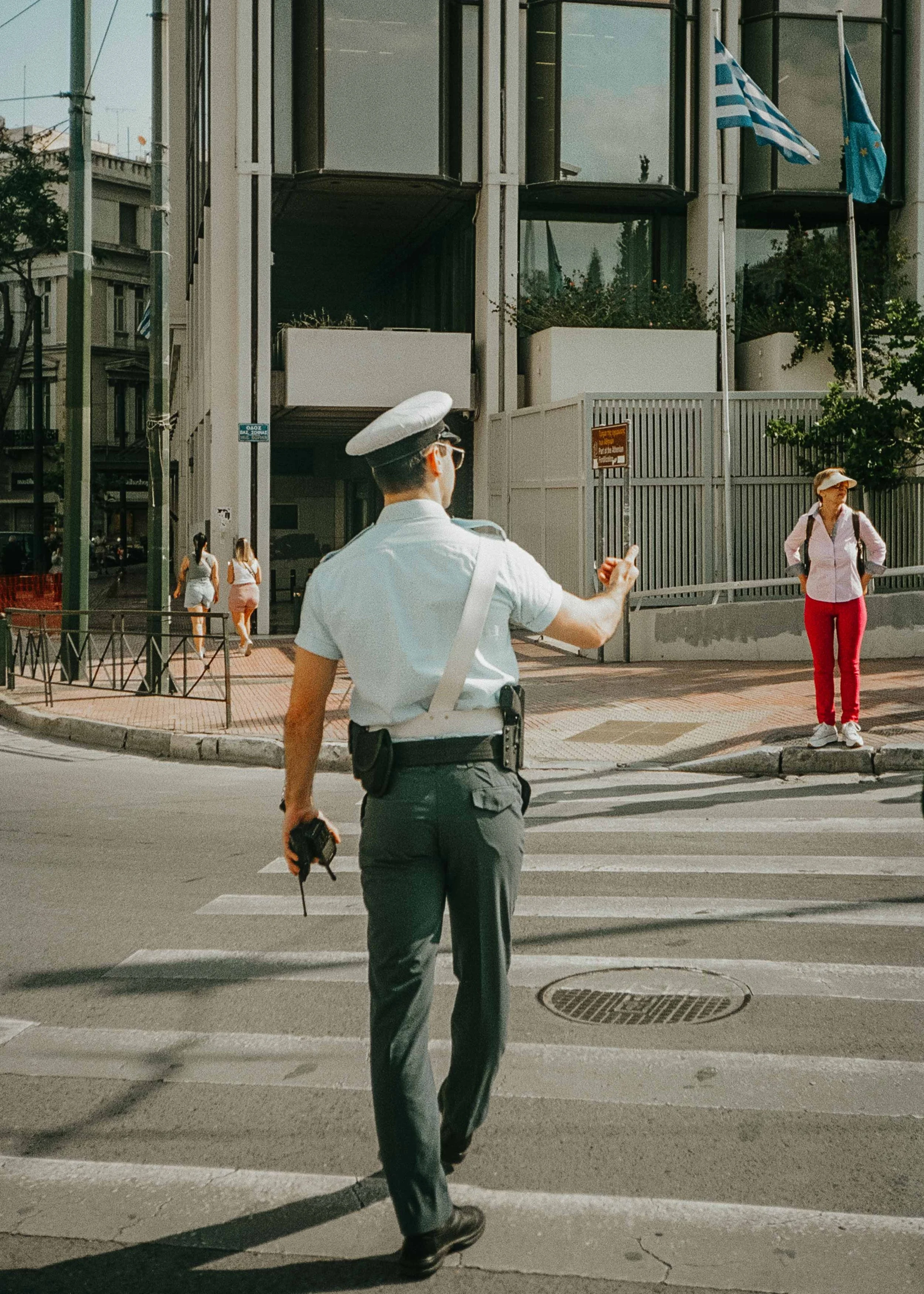 A police officer with a walkie-talkie and a camera crossing a city street while guiding pedestrians and traffic, with two women waiting at the corner, and a modern building with Greek and European Union flags in the background.