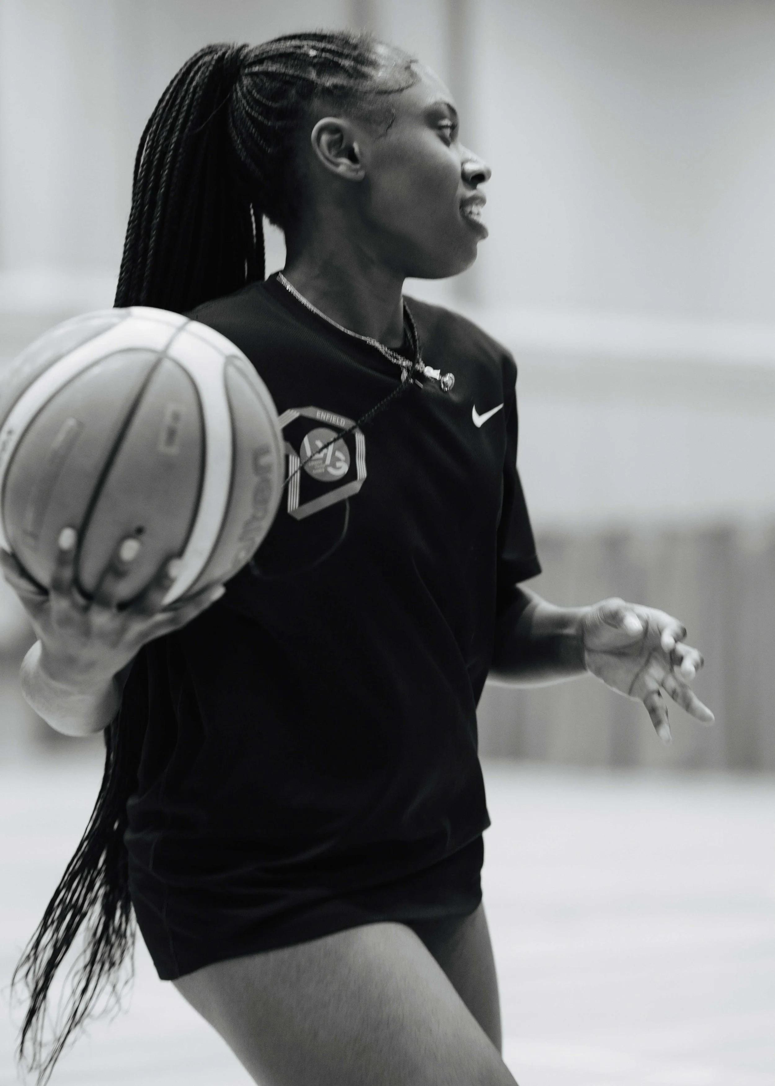 A woman with long braided hair holding a basketball in an indoor gym, wearing a black Nike sports shirt and gray leggings.