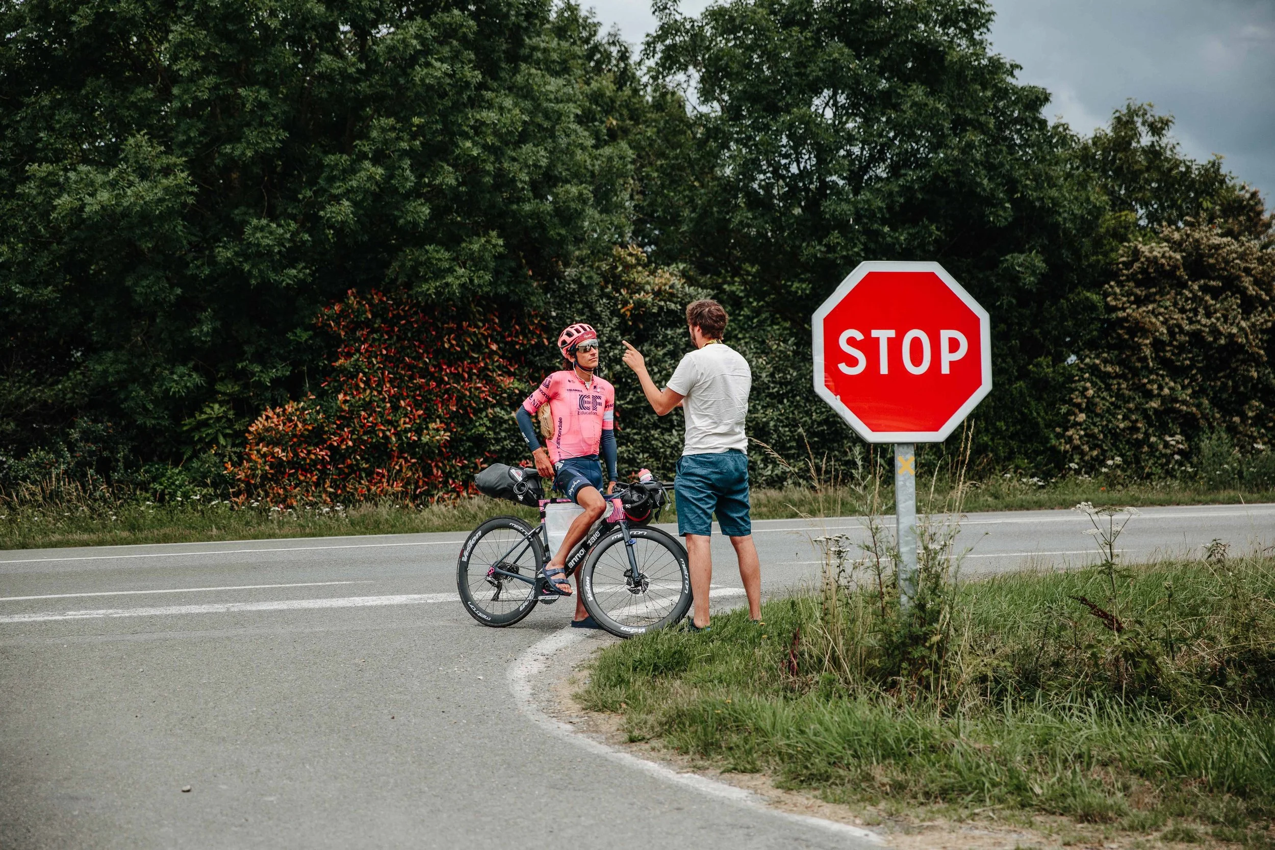 A cyclist in pink riding gear talking to a man in a white shirt and shorts at a stop sign on the side of the road with trees in the background.
