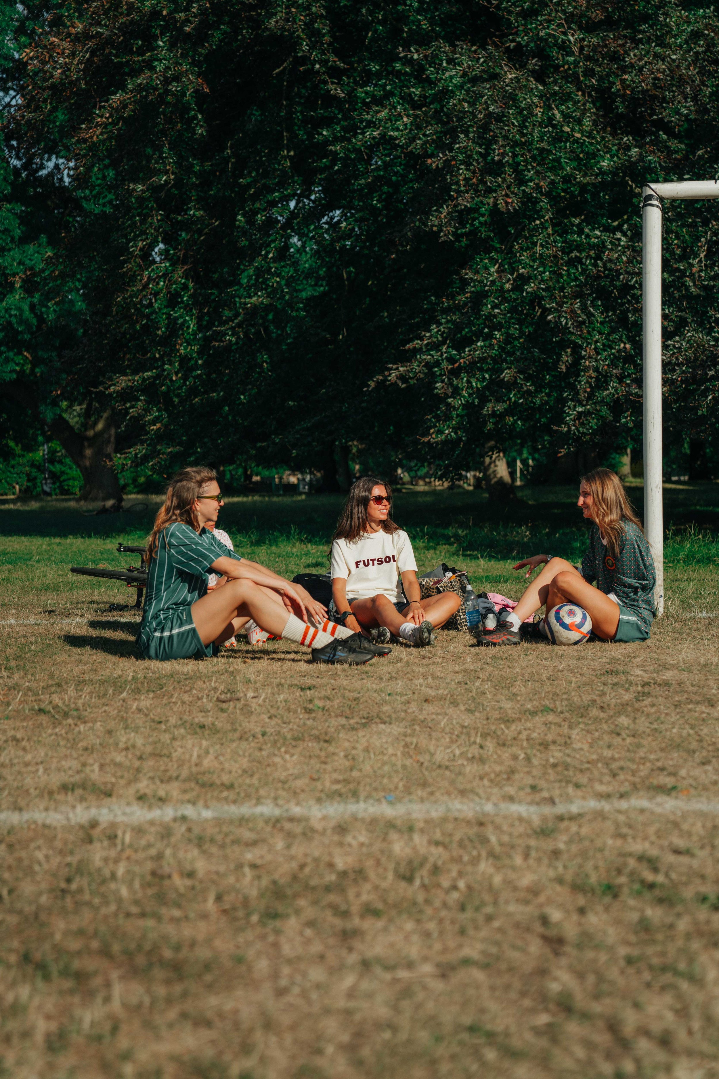 Three young women sitting on the grass near a soccer goal, talking and relaxing during the daytime surrounded by trees.