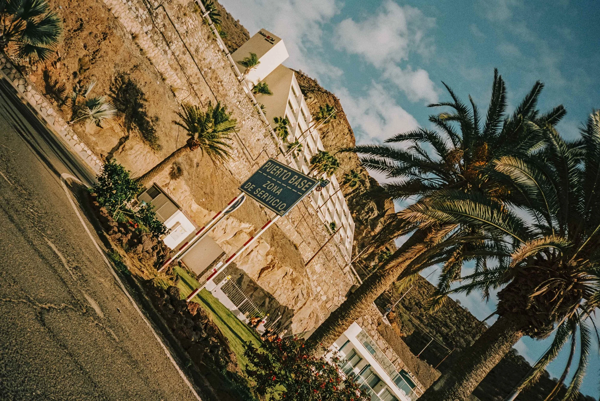 Palm trees next to a blue road sign that reads 'Puerto Base Zona de Servicio,' with white buildings on a hillside and a partly cloudy sky.