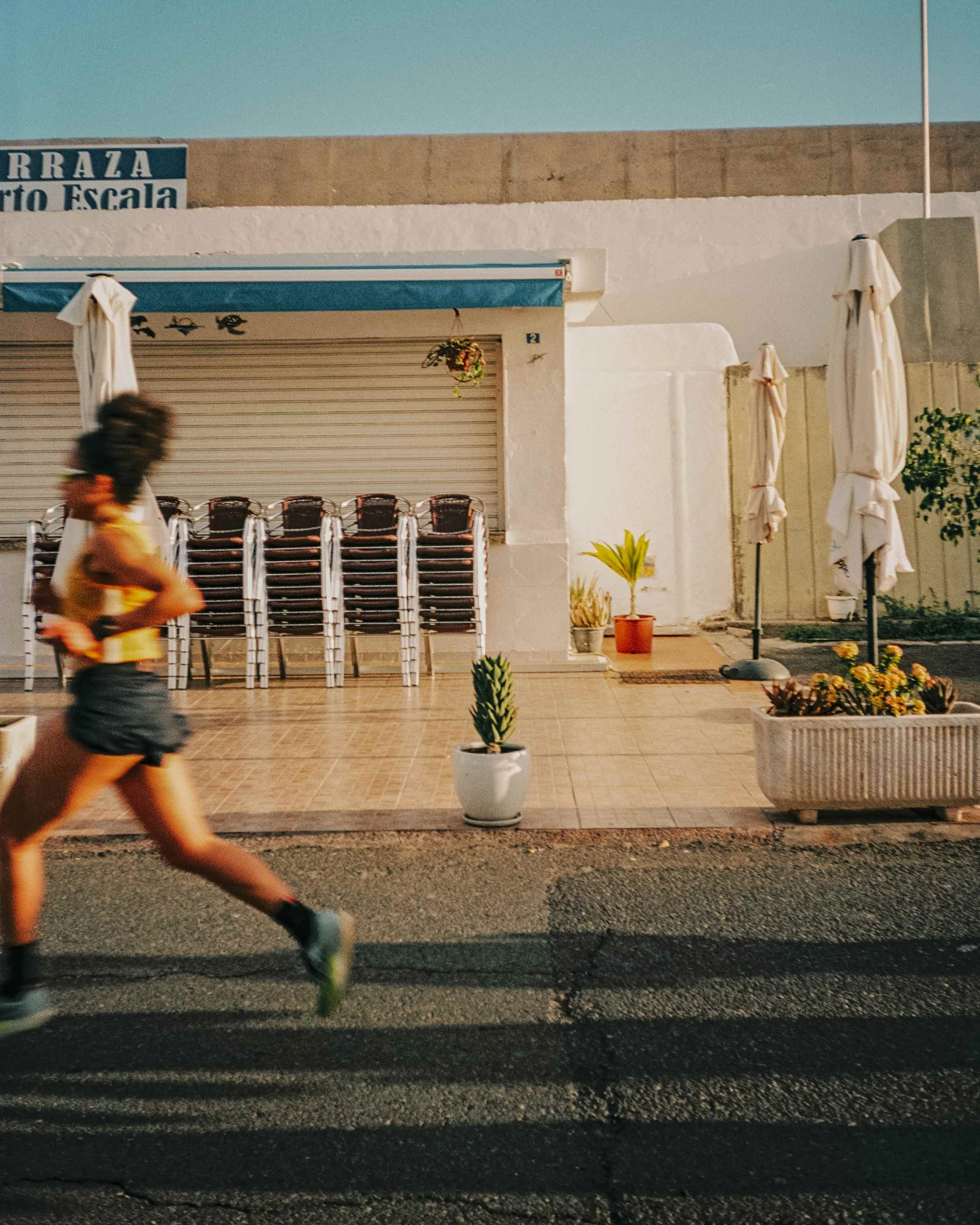 A woman running past a patio with stacked chairs, potted plants, and closed umbrellas outside a building with a sign in Spanish.