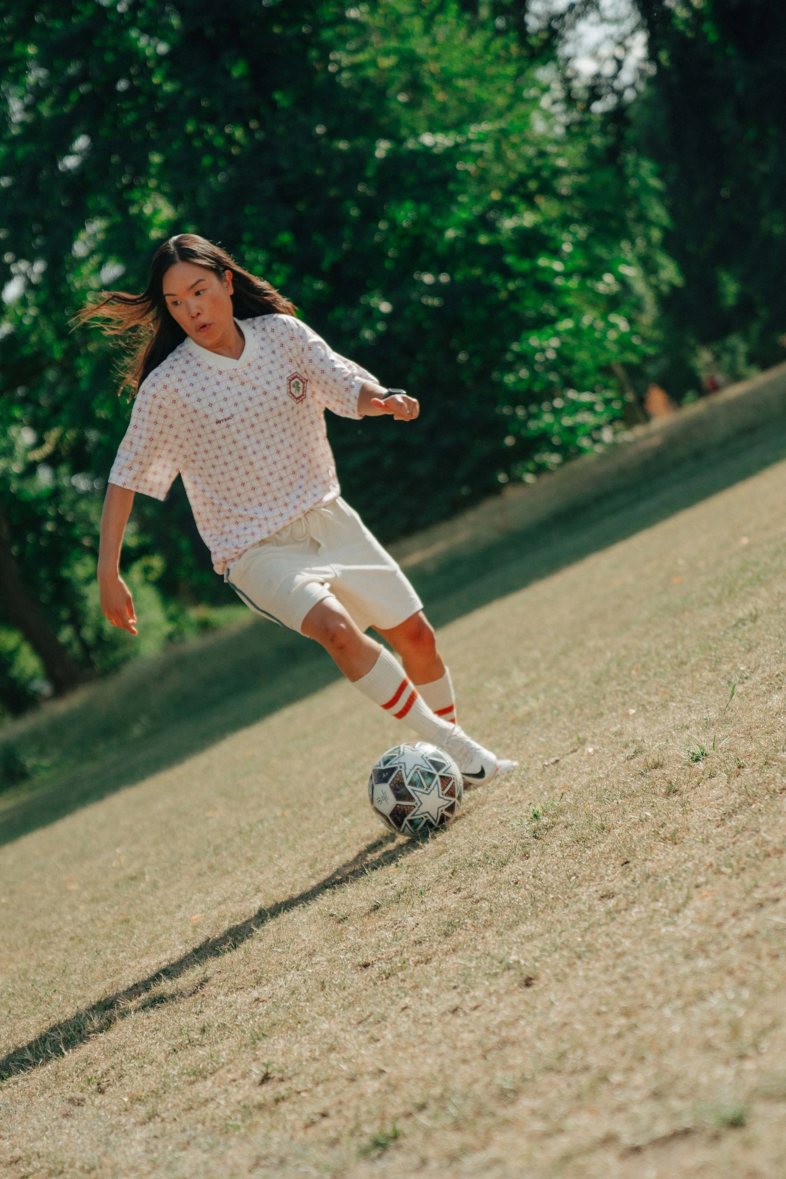 A young girl playing soccer outdoors, kicking a ball on a grassy field with trees in the background.