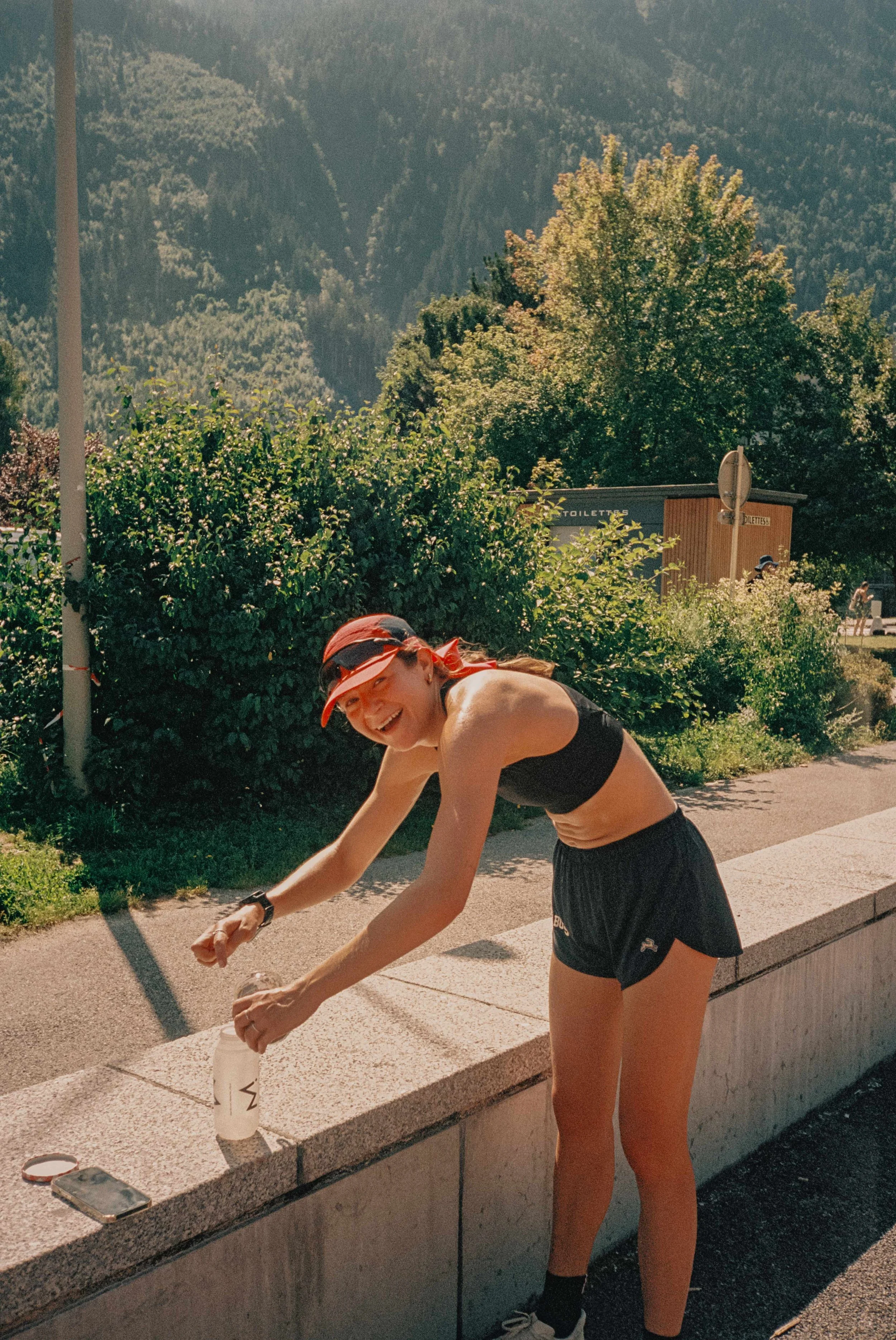 A woman in athletic wear and a red visor pouring water from a bottle onto her face outdoors with mountains and greenery in the background.