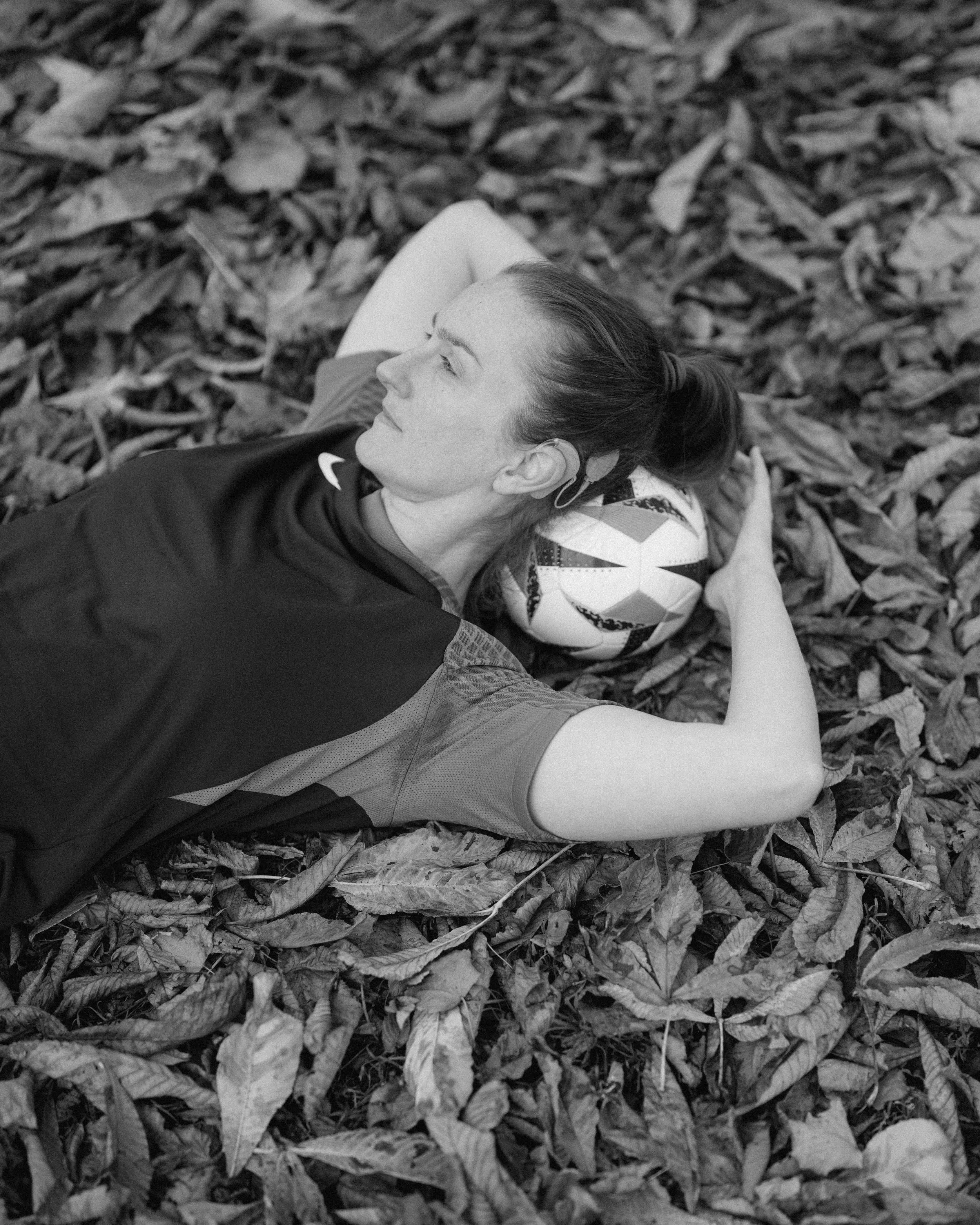 A woman lying on her side on a bed of fallen leaves, resting her head on a soccer ball, outdoors.