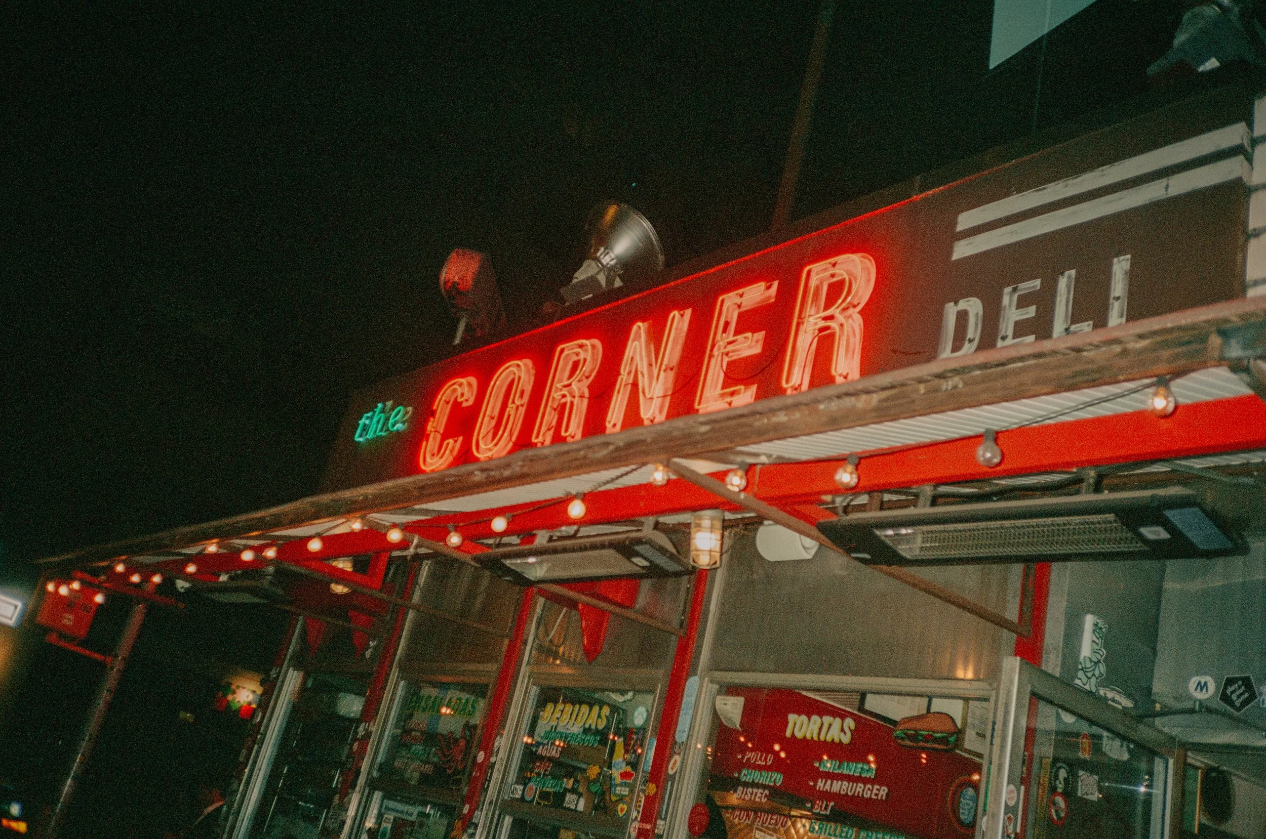 Night view of a restaurant with a red neon sign reading 'The Corner Deli' and string lights above the entrance.