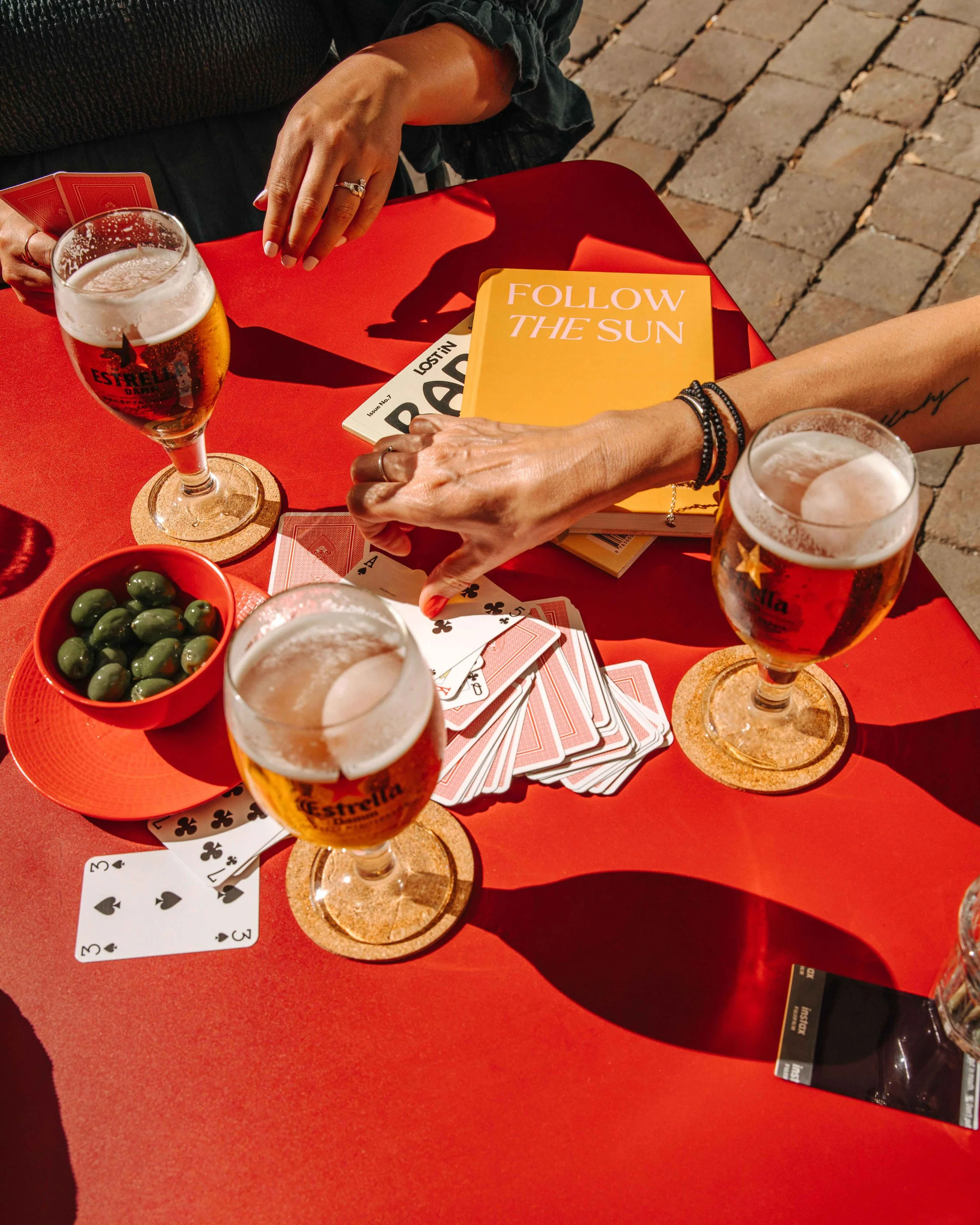People playing cards at a red outdoor table with glasses of beer, a bowl of green olives, and a magazine titled "Follow the Sun" displayed.