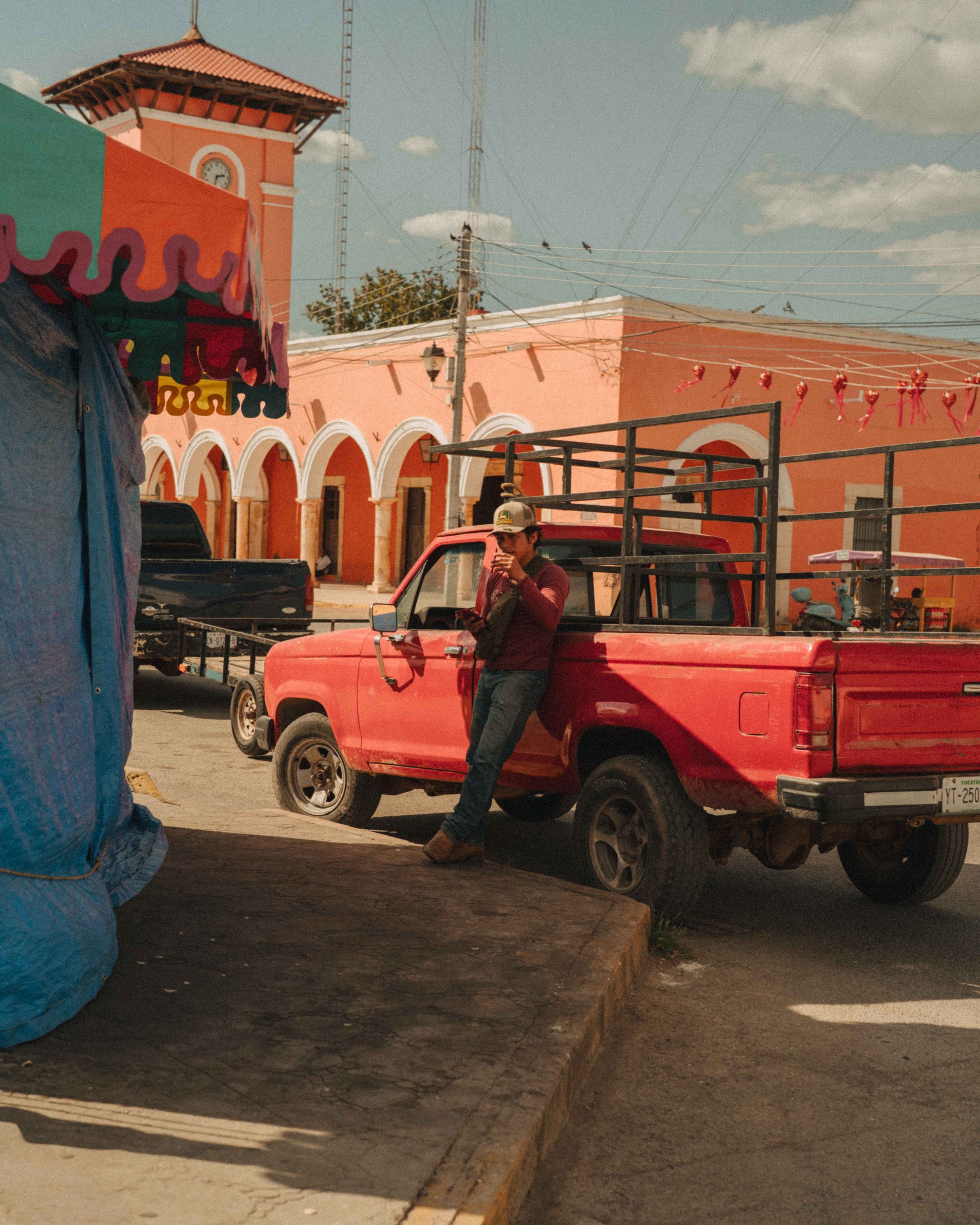 A young man leaning against a red pickup truck, holding a smartphone, in a colorful town square with pink and orange buildings, power lines, and a clock tower in the background.