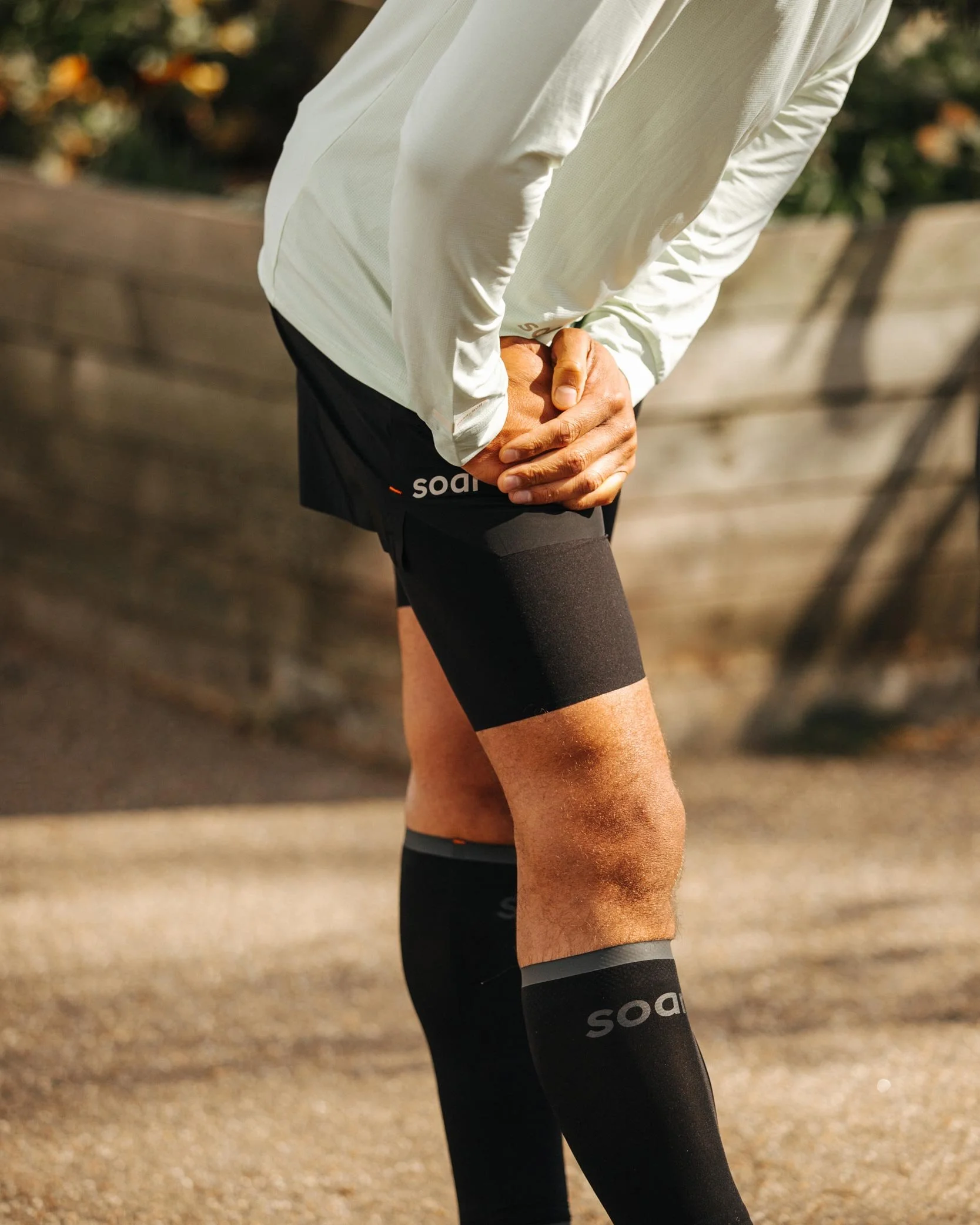 Person clutching their right knee outdoors, wearing black compression socks and athletic shorts, possibly experiencing discomfort.