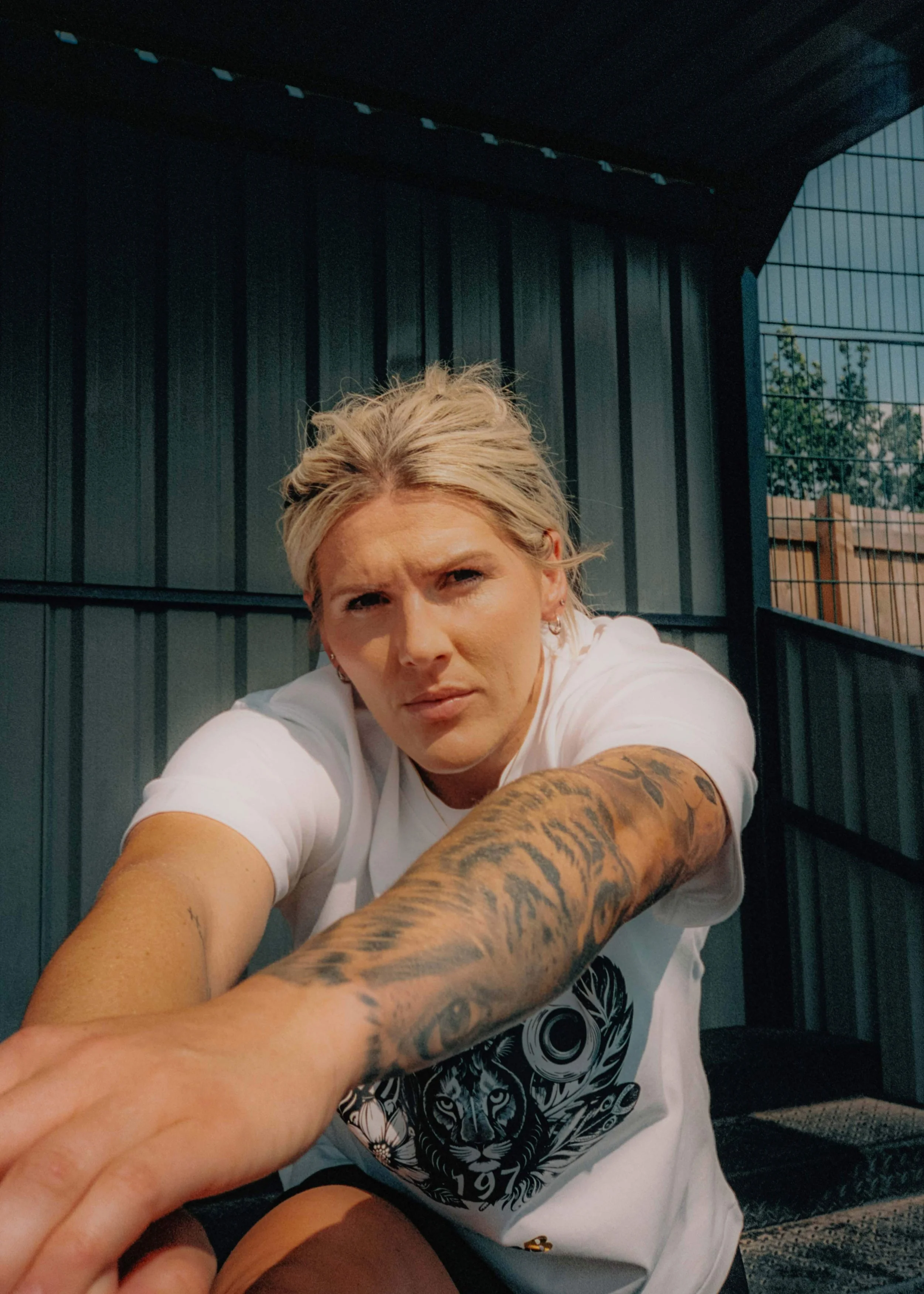 A woman with short blonde hair and tattoos on her arm, wearing a white t-shirt with a black graphic, taking a selfie outdoors against a metal fence.