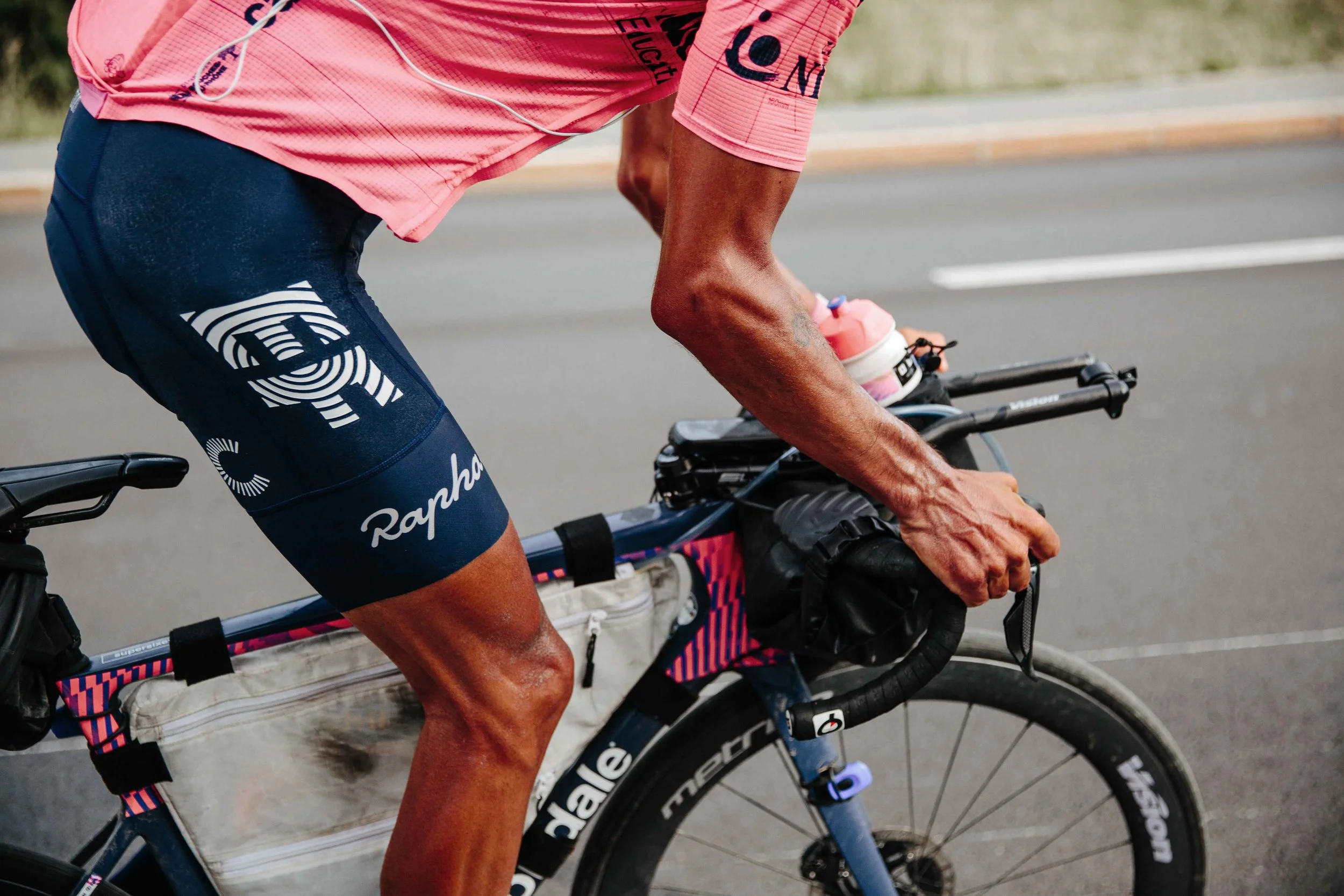 A cyclist wearing pink jersey and dark shorts is riding a mountain bike on a paved road, holding the handlebar with both hands, with a water bottle on top of the handlebar.
