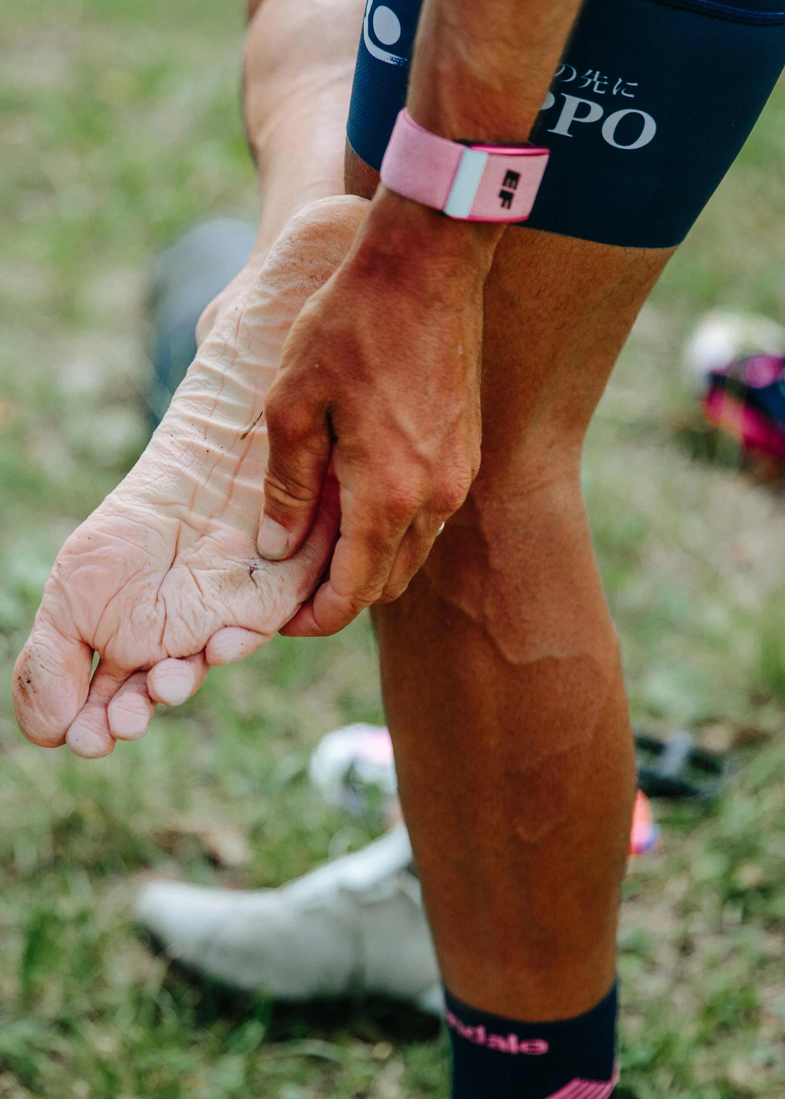 A person in athletic gear stretching outdoors, touching the ground with one hand, with a focus on their tanned legs and wrinkled hand.