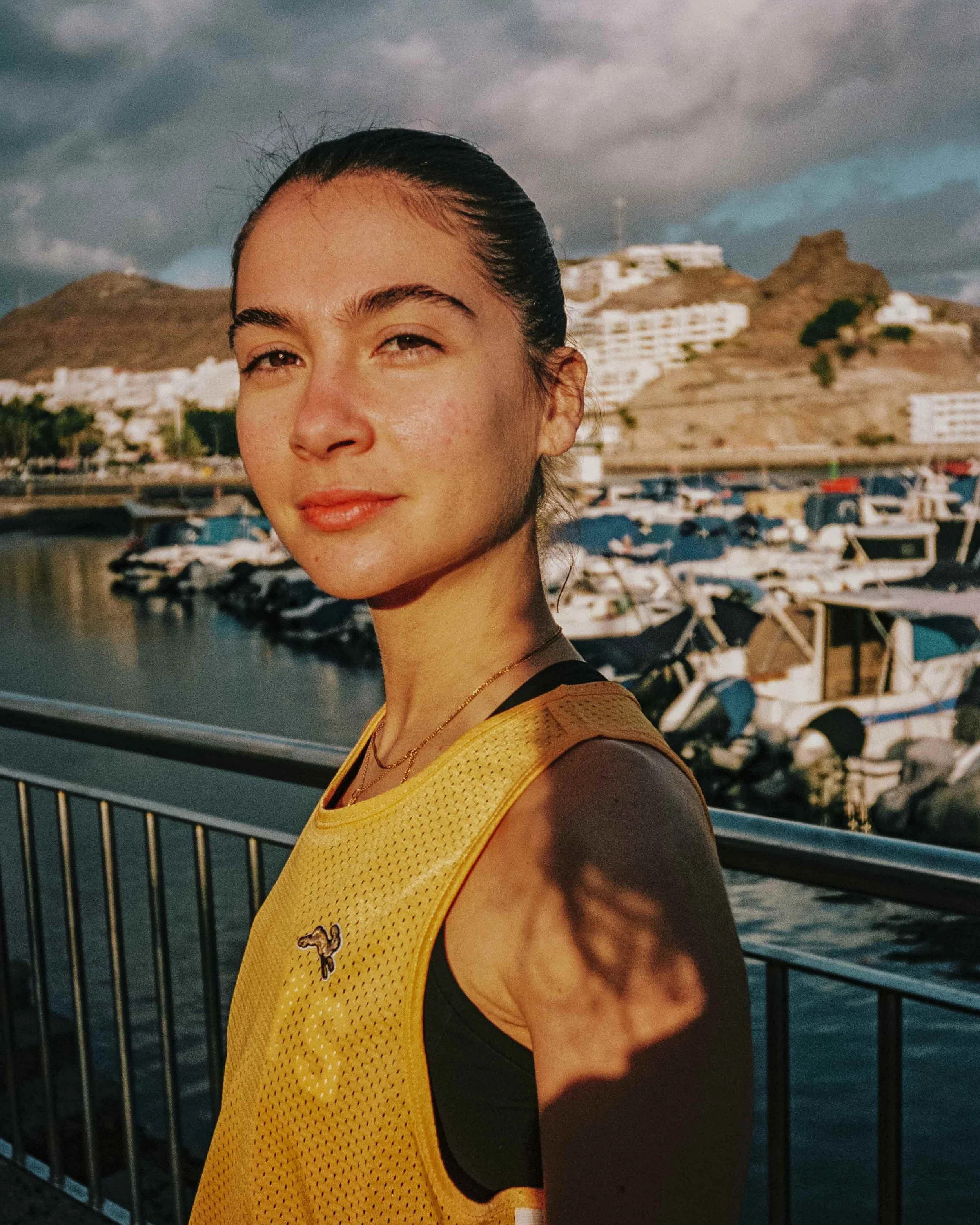 A young woman with dark hair tied back, wearing a yellow sports tank top, standing outdoors near a marina with boats docked, hills and white buildings in the background, under a cloudy sky.