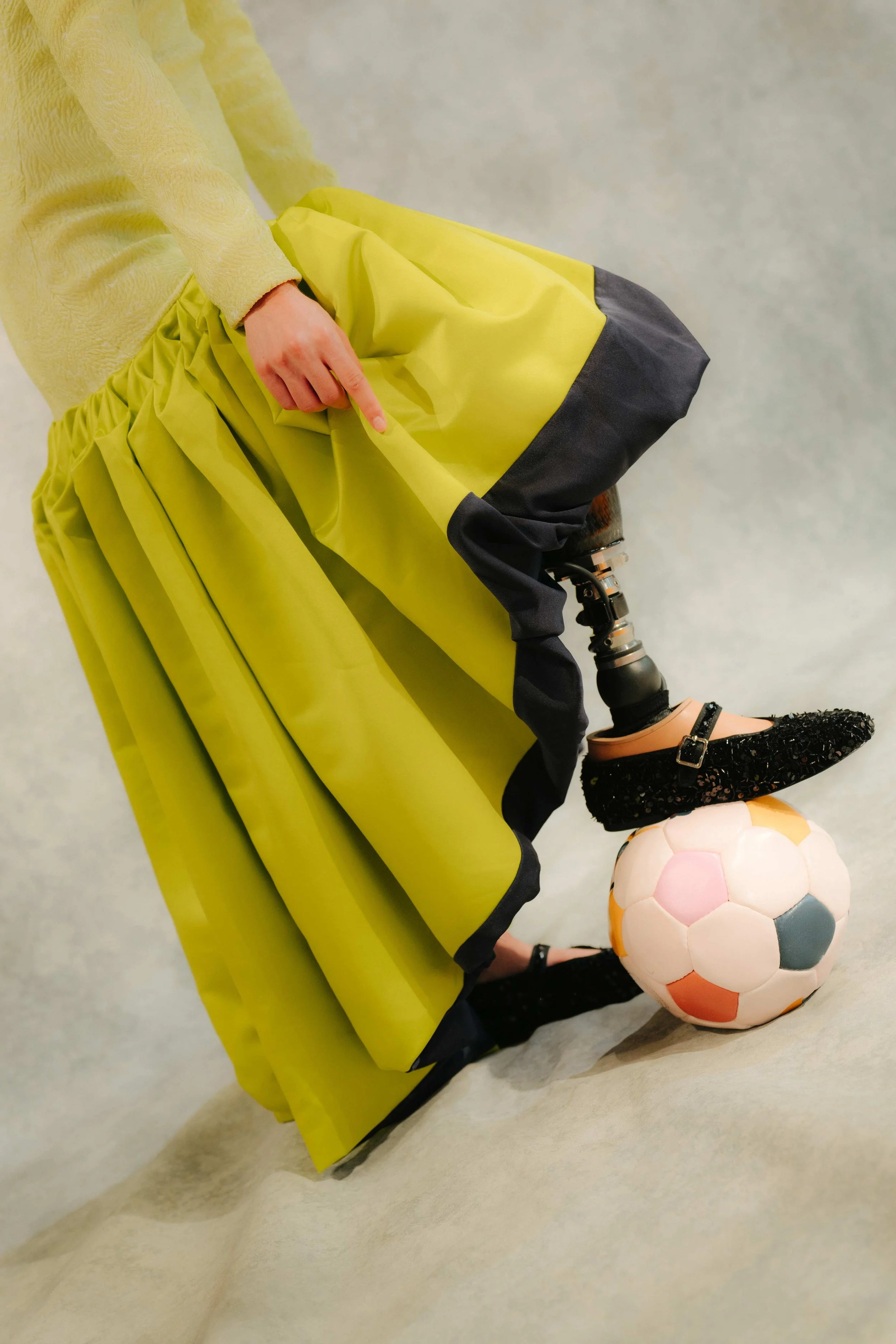 Close-up of a prosthetic leg with a black sneaker on a colorful soccer ball. The person is wearing a yellow and black skirt and a yellow textured sweater.