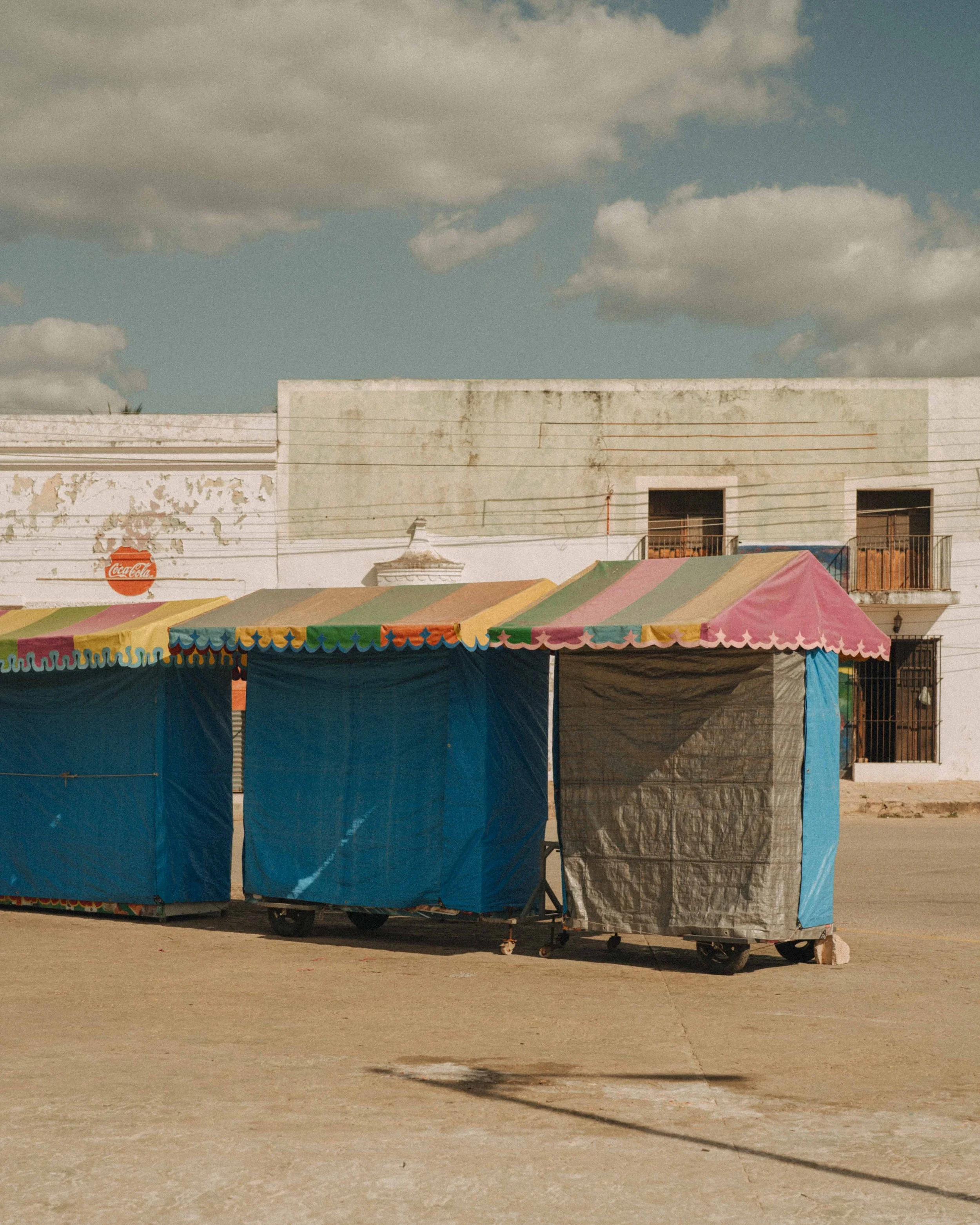 Colorful tents with striped tops set up outdoors in a deserted area with a white building in the background, under a sky with scattered clouds.