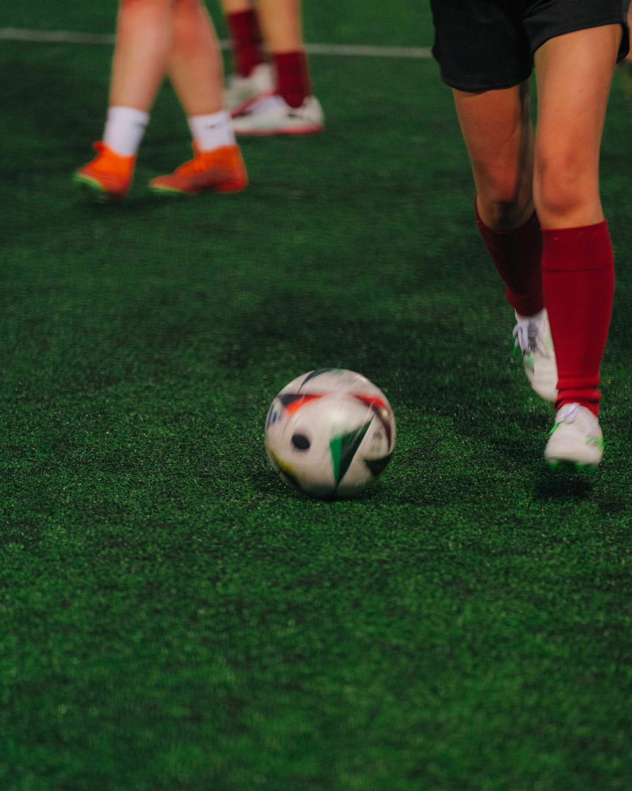 Close-up of a soccer ball on artificial turf with players in socks and shoes in the background.