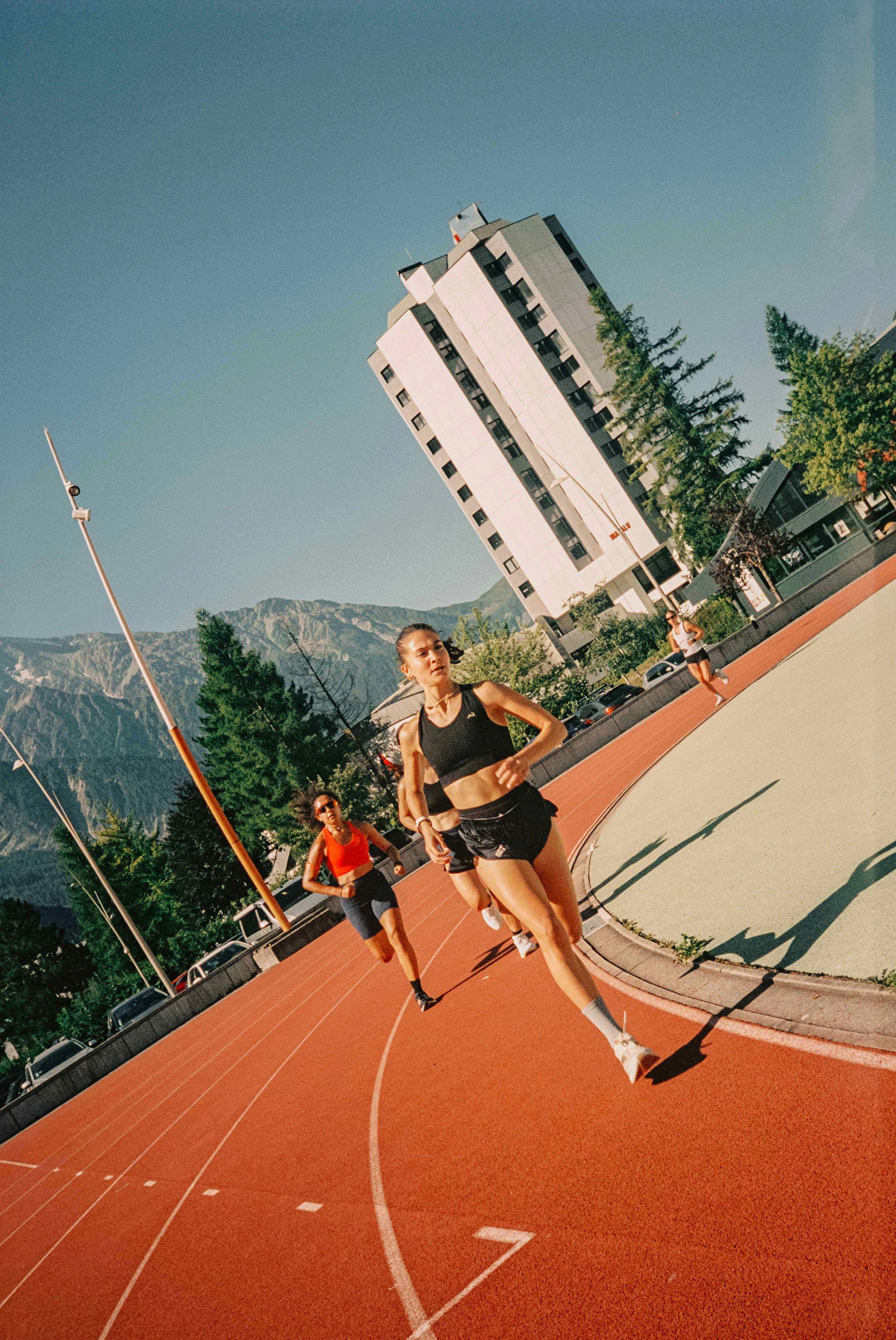 Three women running on a red outdoor track during daytime with mountains and a tall white building in the background.