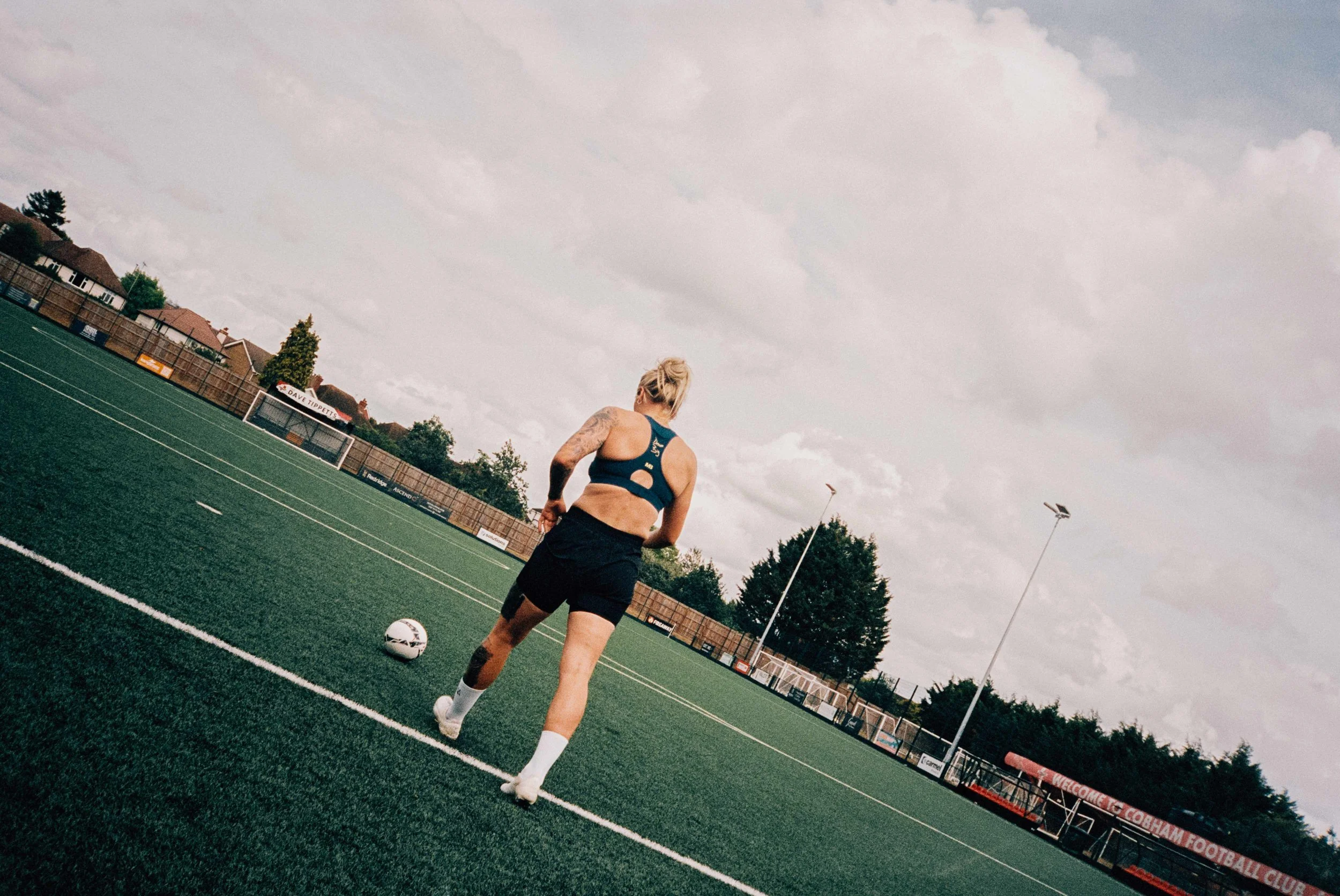 A female soccer player with tattoos, wearing black shorts and a blue sports bra, is running on a football field with a soccer ball near her feet.