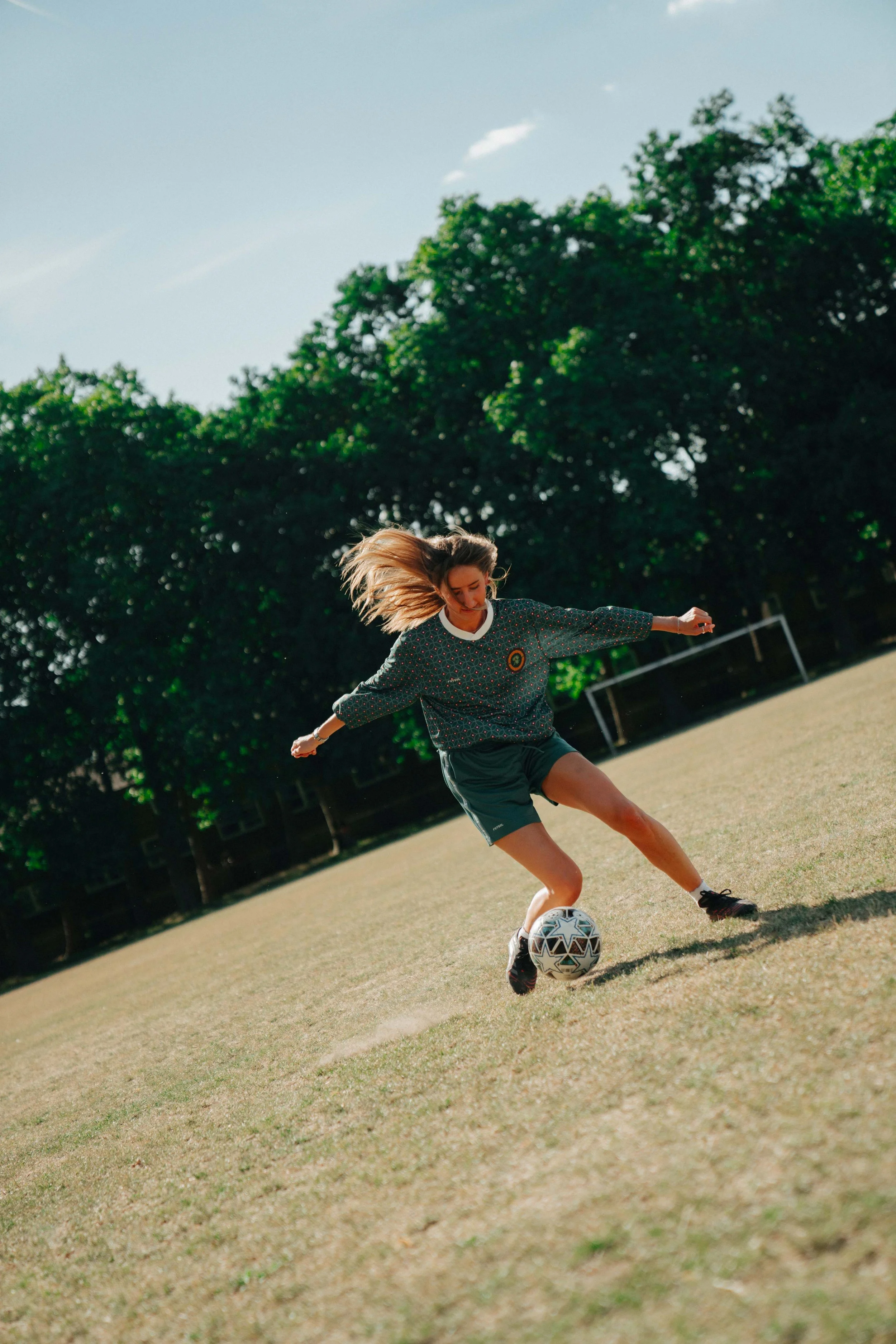 Young woman playing soccer on a grassy field with trees and a cloudy sky in the background.
