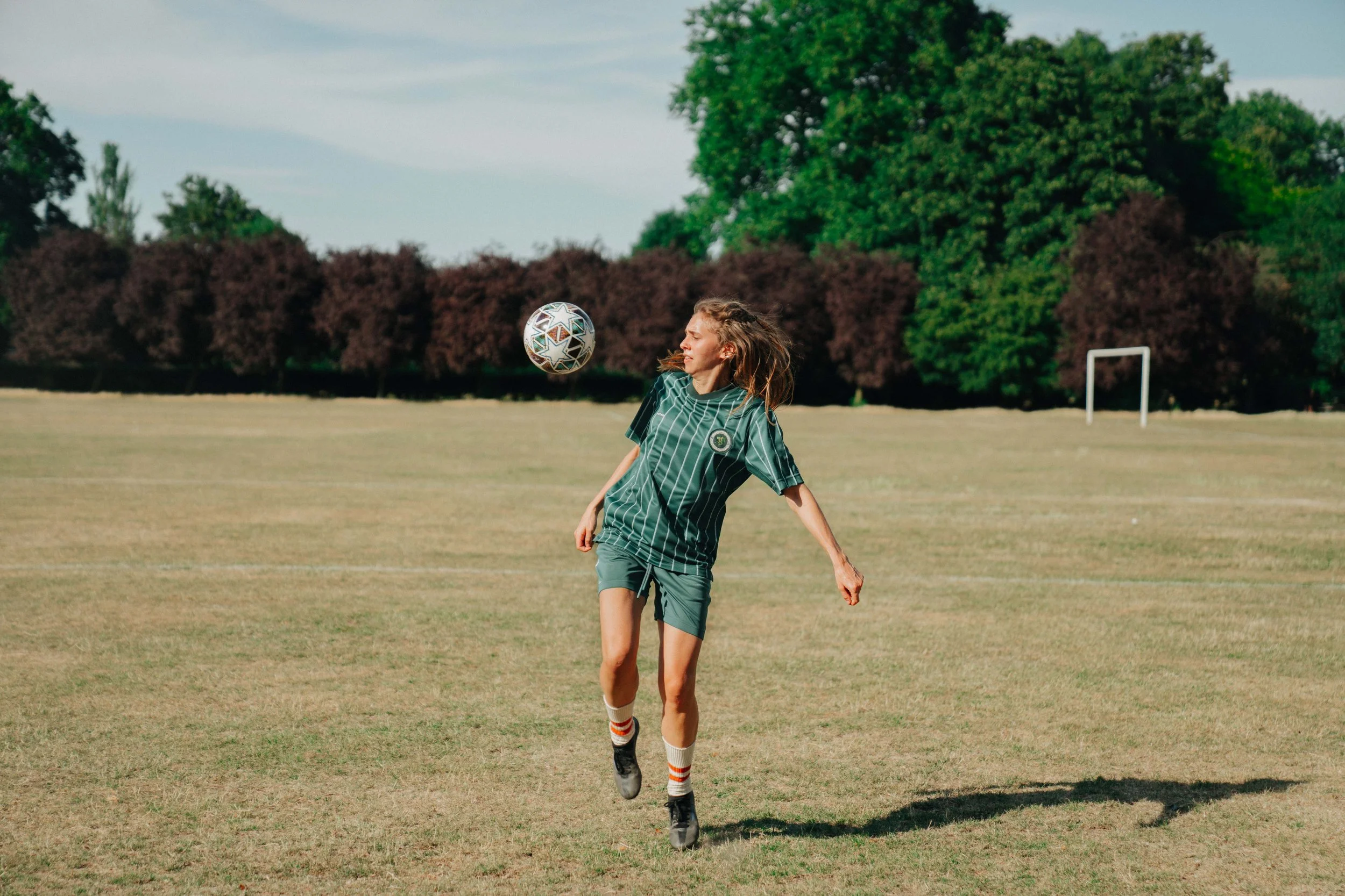 A girl in a green sports uniform playing soccer on a grassy field with trees in the background.