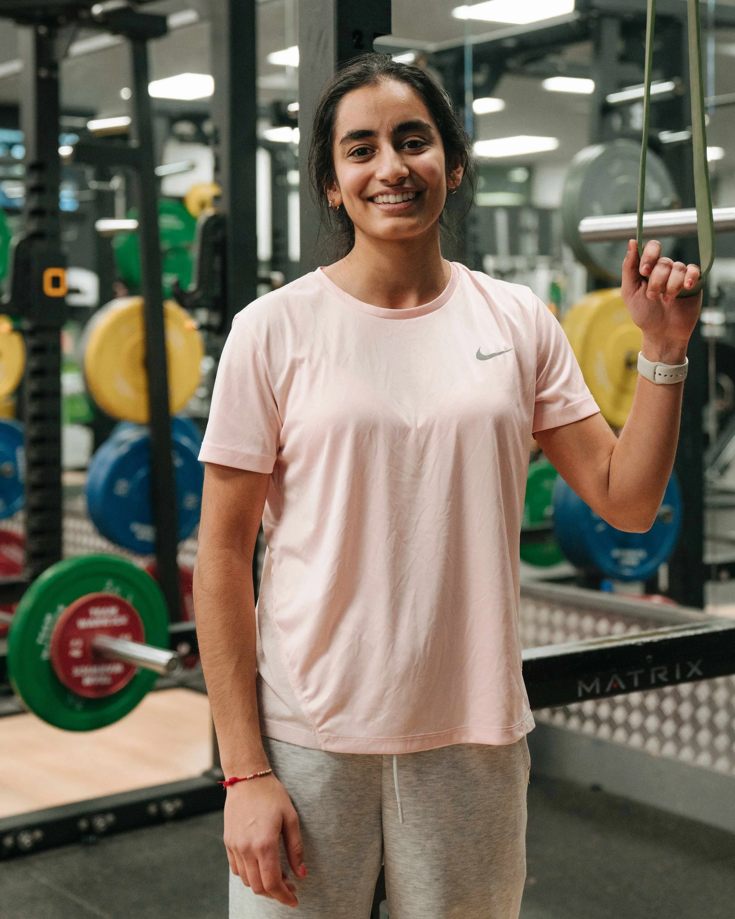 Young woman smiling at the gym, holding a resistance band, in casual workout attire, with weightlifting equipment in the background.