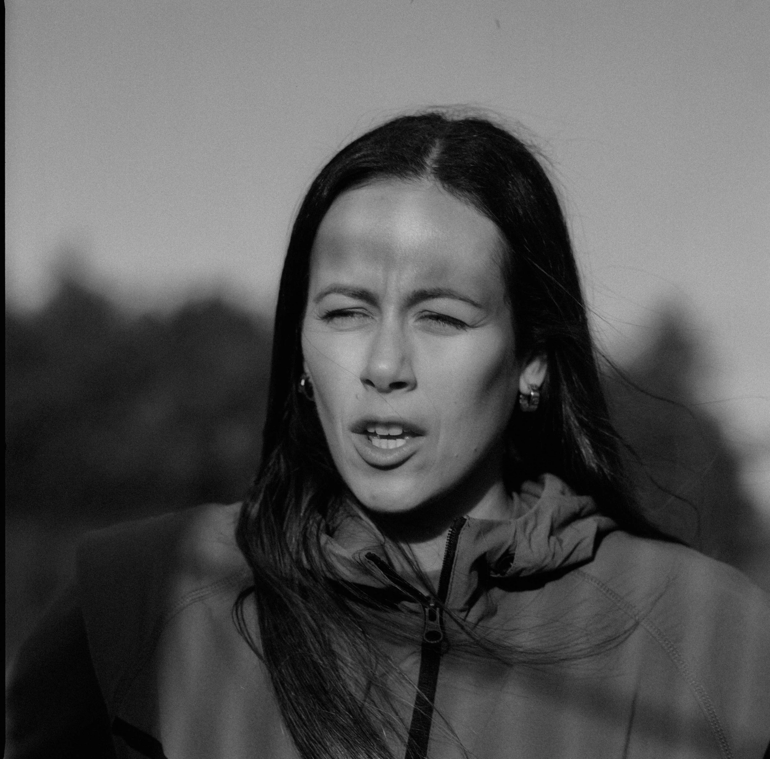 Black and white photo of a woman with long dark hair, squinting and slightly opening her mouth, wearing earrings and a jacket with a high collar outdoors.