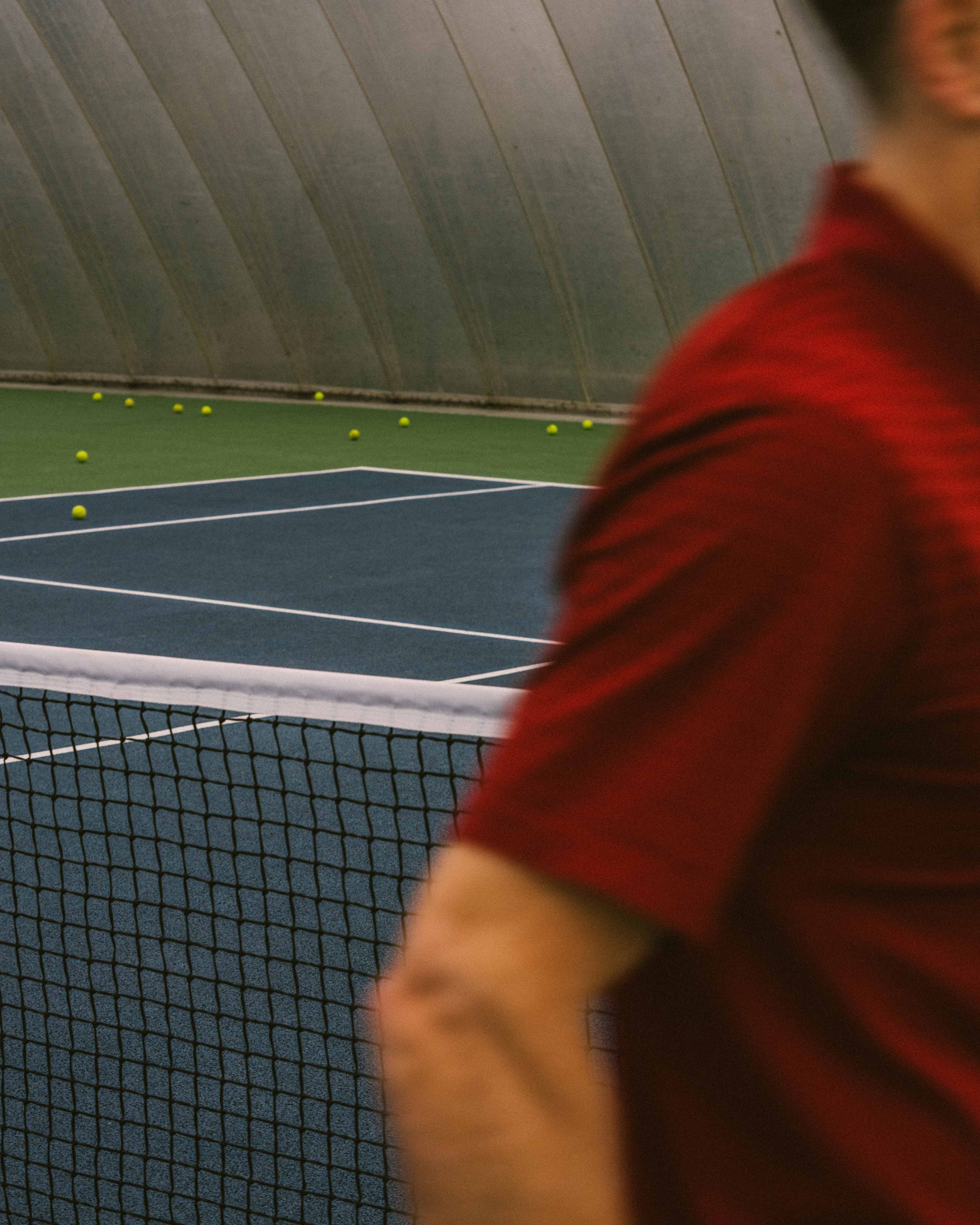 Part of a tennis court with yellow tennis balls scattered around, a black net, and a person in a red shirt standing near the net.