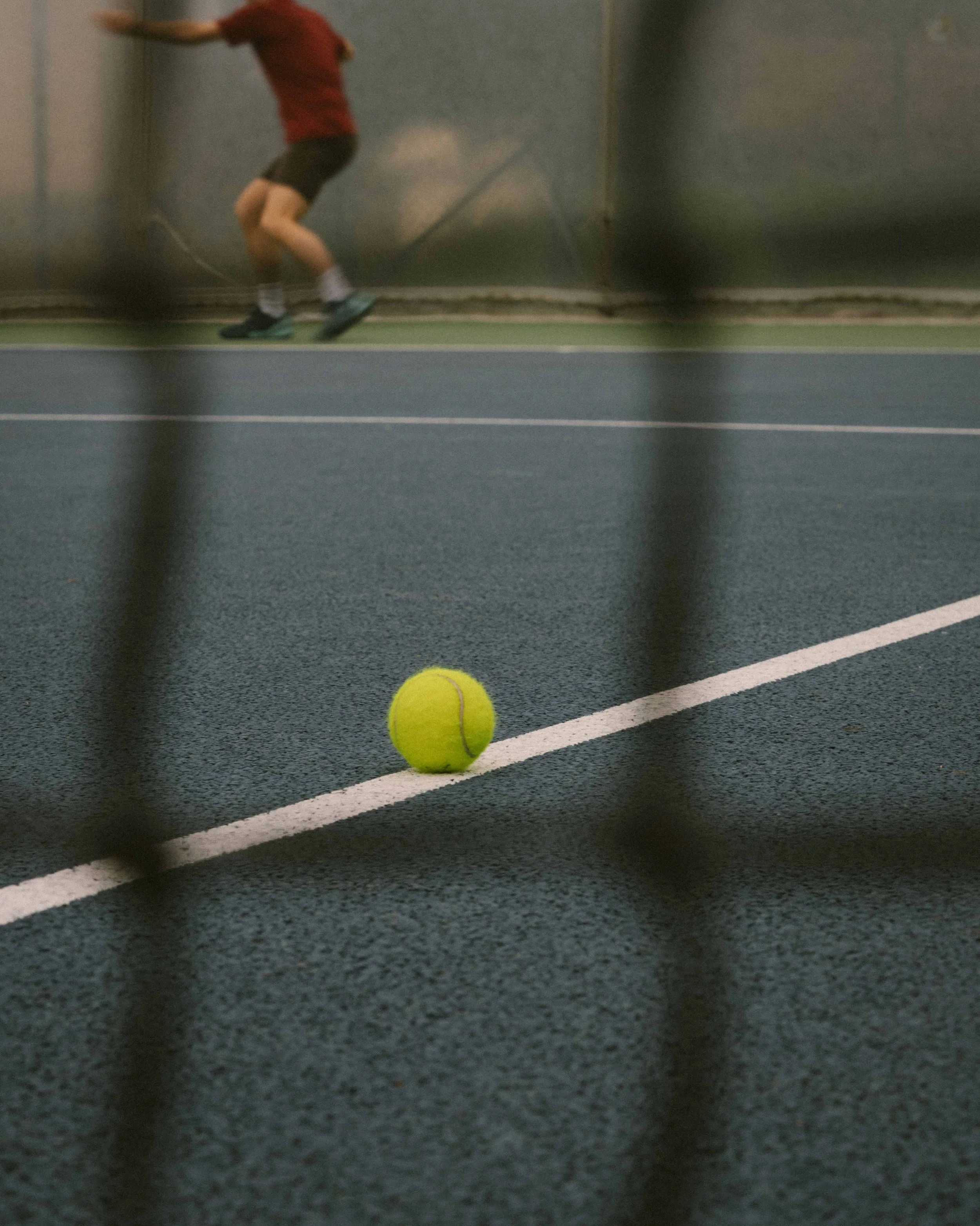A tennis ball on a court, seen through a chain-link fence, with a person in a red shirt and dark shorts in the background.