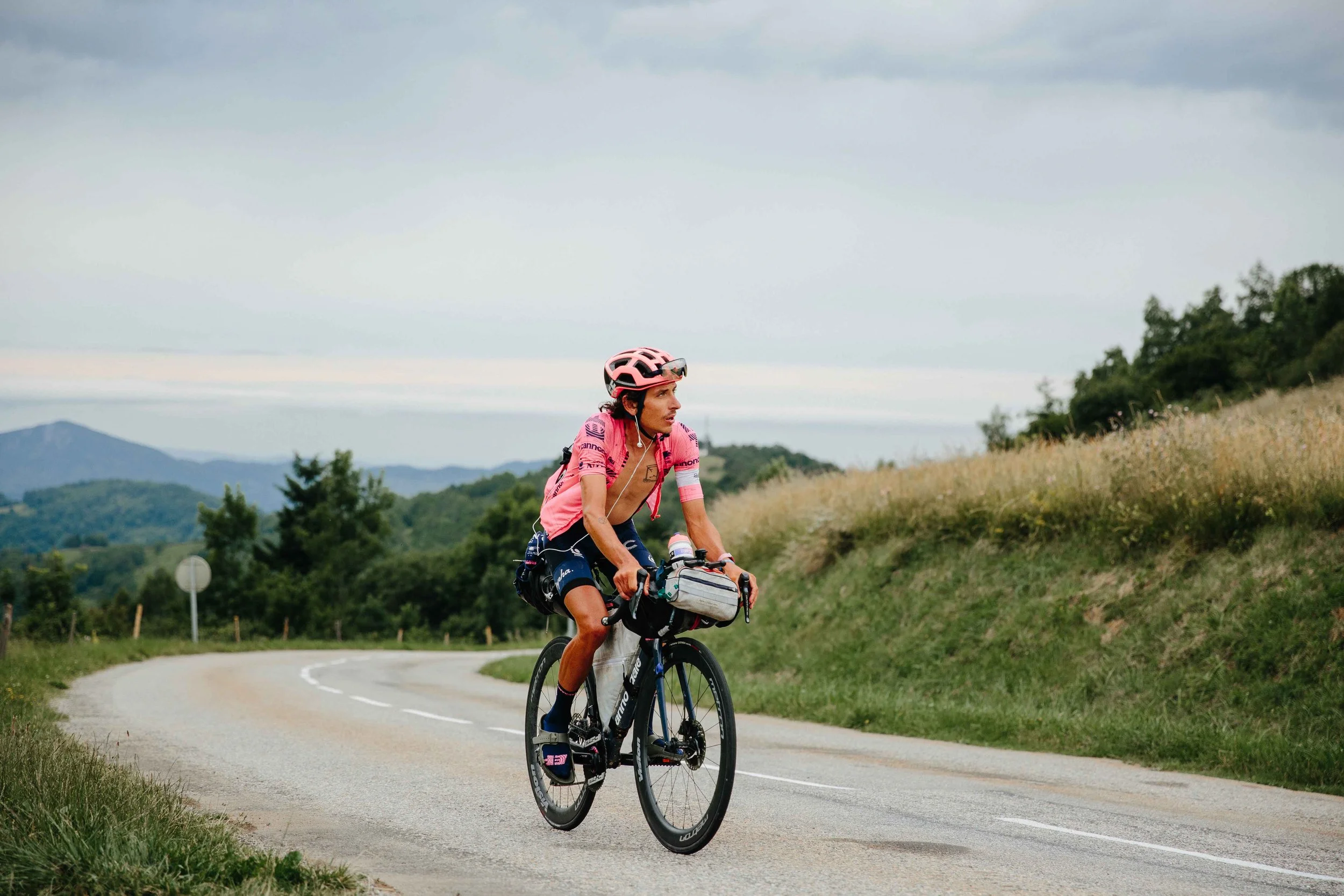 A cyclist wearing a pink helmet and jersey riding a mountain bike on a winding rural road with grass and trees on either side, under a cloudy sky.